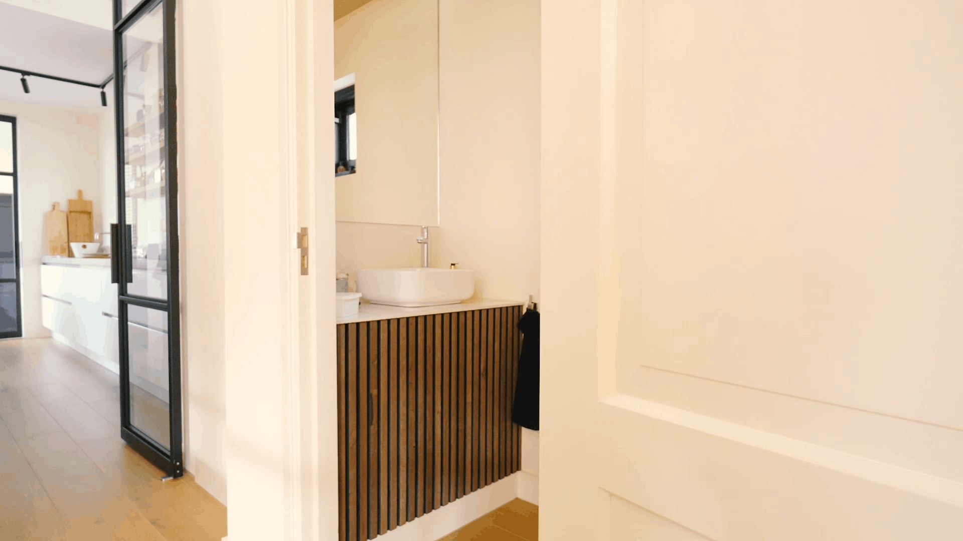 Bagno moderno con lavabo rotondo, mobile acustico a doghe di legno, grande specchio e porta bianca accanto a un luminoso corridoio e a una cucina contemporanea.