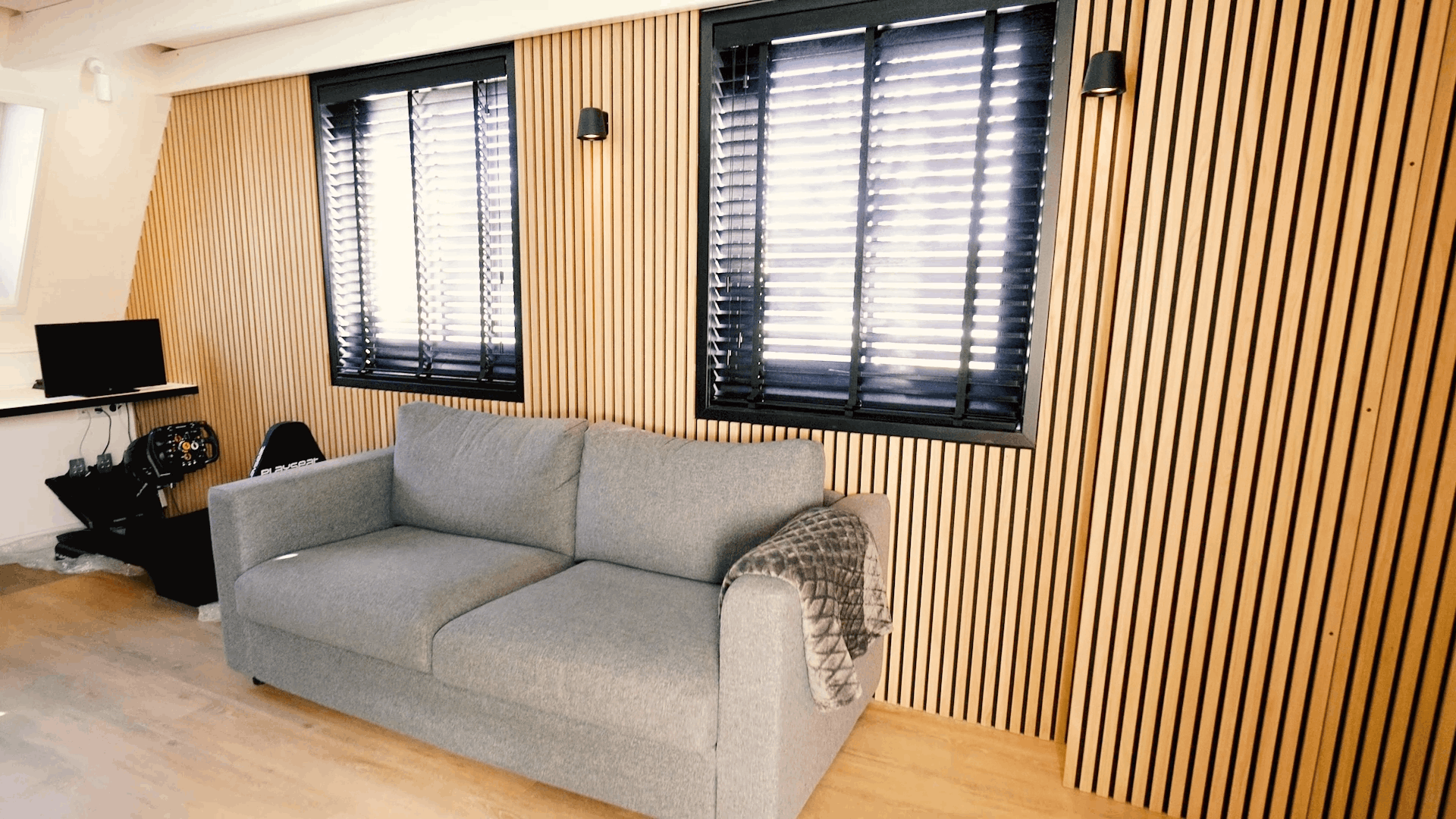 Divano grigio con coperta a fantasia davanti a una parete di doghe acustiche in legno Akupanel nordico, affiancata da due finestre con tende nere e da una scrivania compatta.