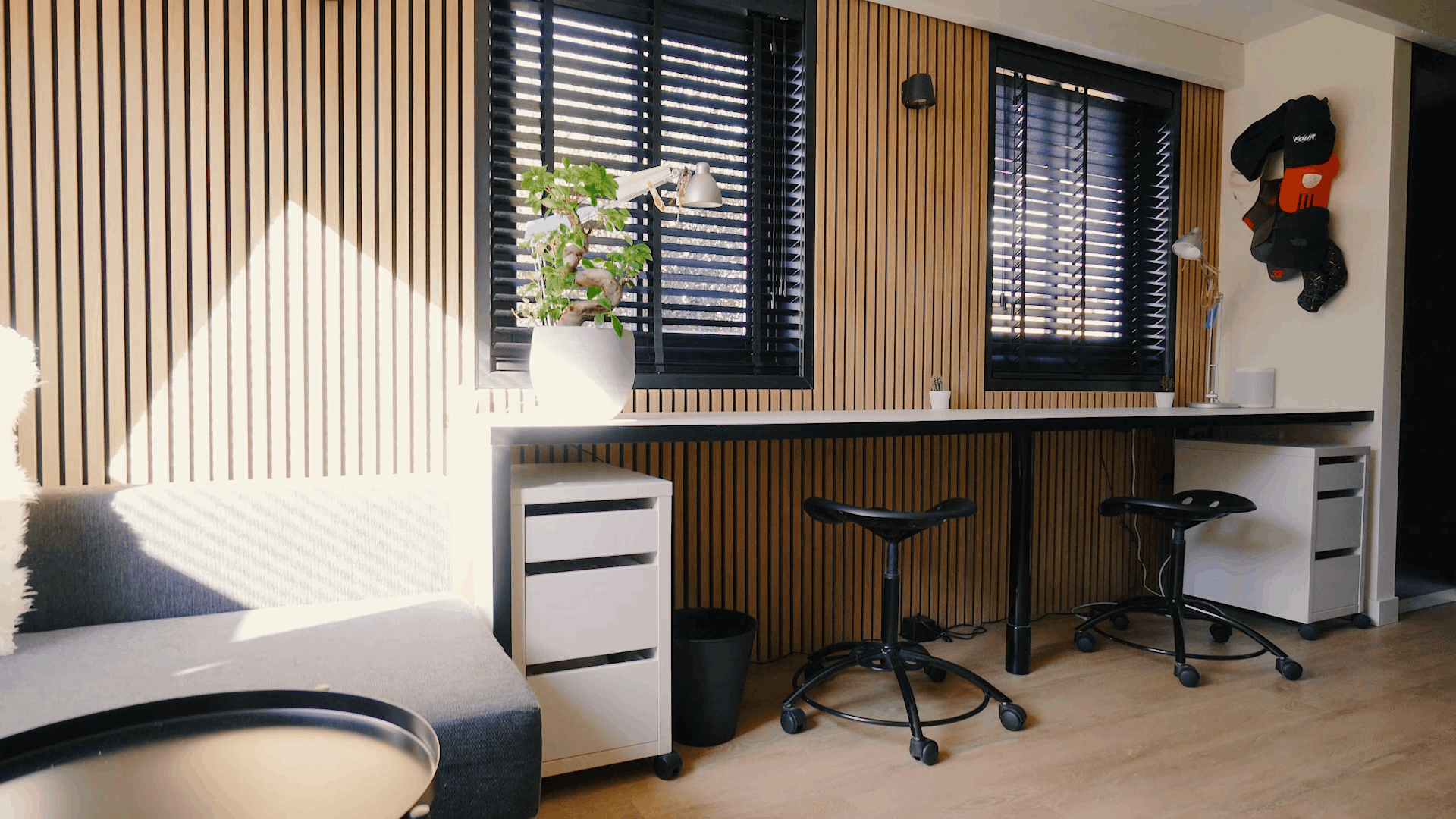 Ufficio moderno con pannelli in legno acustico nordico alle pareti, postazioni di lavoro ergonomiche, sgabelli neri, una pianta in vaso e la luce del sole attraverso le tende nere della finestra.