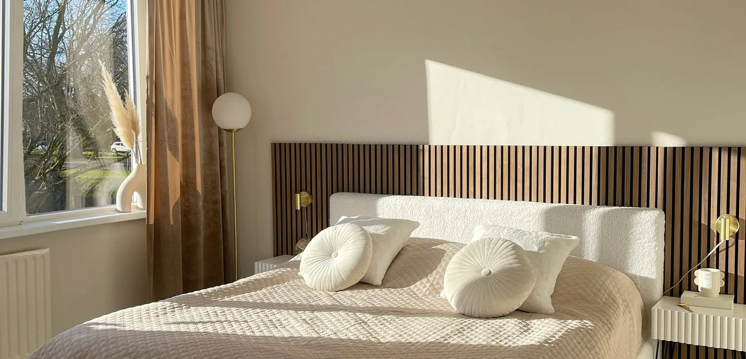 Camera da letto moderna illuminata dal sole con letto trapuntato beige, cuscini rotondi, testiera a doghe in legno Akupanel e arredamento minimal accanto a una grande finestra con tende trasparenti.