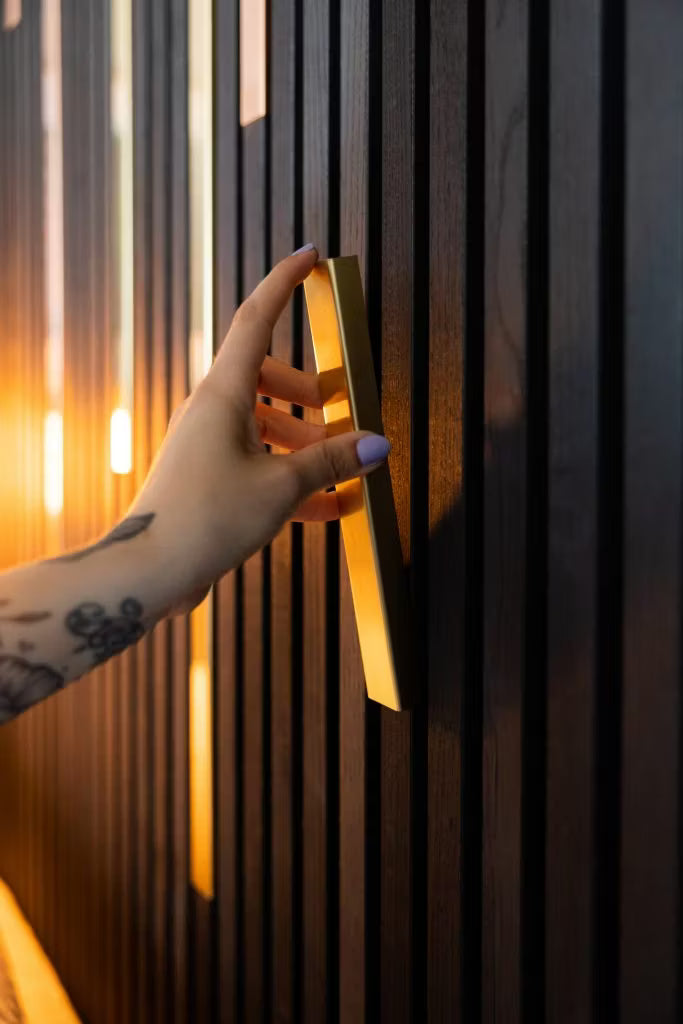 Una mano tatuata con unghie viola chiaro stringe una maniglia doro su una parete elegante e scura in legno acustico Akupanel, sottolineata da unilluminazione calda.