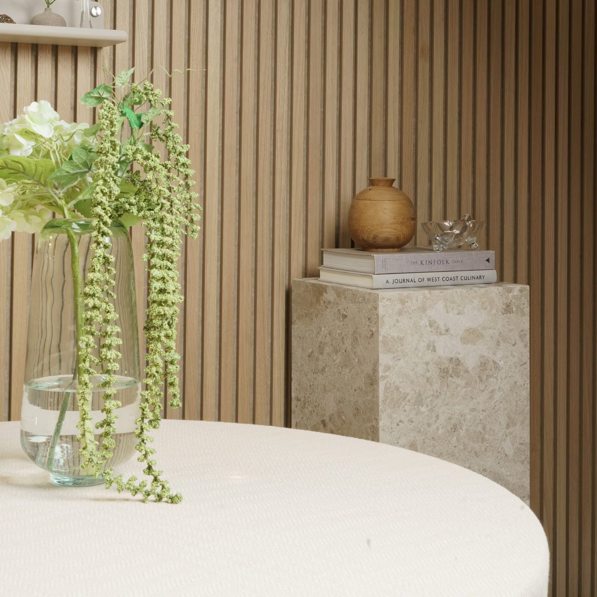 Un vaso di fiori si trova su un tavolo rotondo; libri e ceramiche sono appoggiati su un piedistallo di marmo contro una parete in pannelli di legno acustico Akupanel.