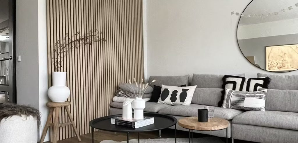 Soggiorno moderno con divano grigio, cuscini astratti, tavolini neri, vasi decorativi e parete acustica in legno Akupanel con specchio rotondo.