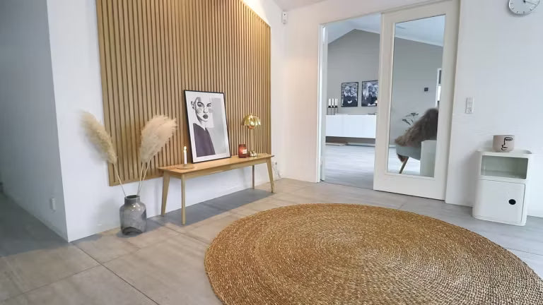 Soggiorno nordico minimalista con tappeto rotondo in iuta, panca in legno con erba della pampa, opere darte incorniciate e parete daccento in legno acustico Akupanel a doghe.