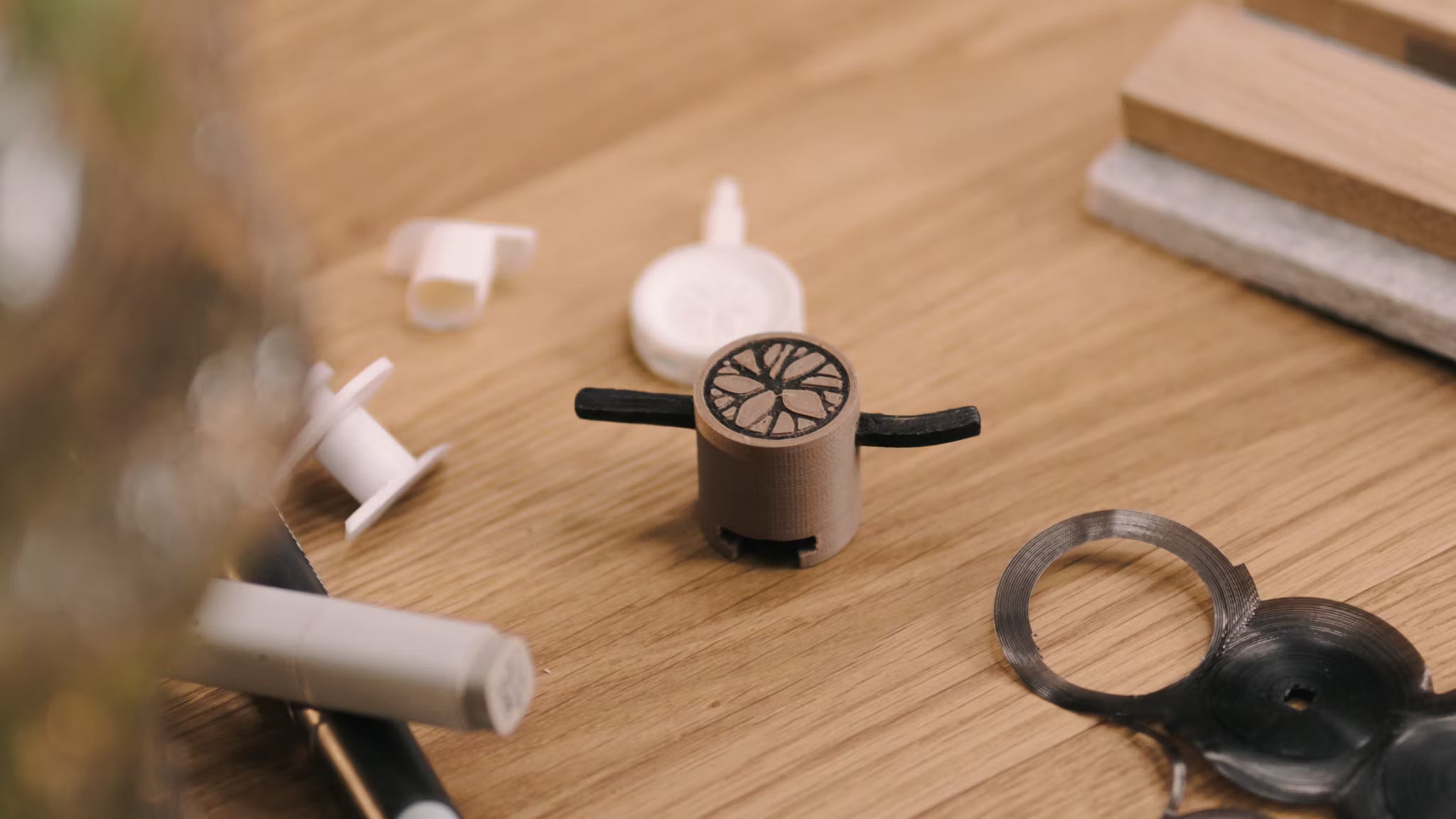 Un piccolo prototipo stampato in 3D con braccia nere si trova su una scrivania di legno accanto a vari componenti in plastica, facendo riferimento allo sviluppo di accessori per pannelli acustici.
