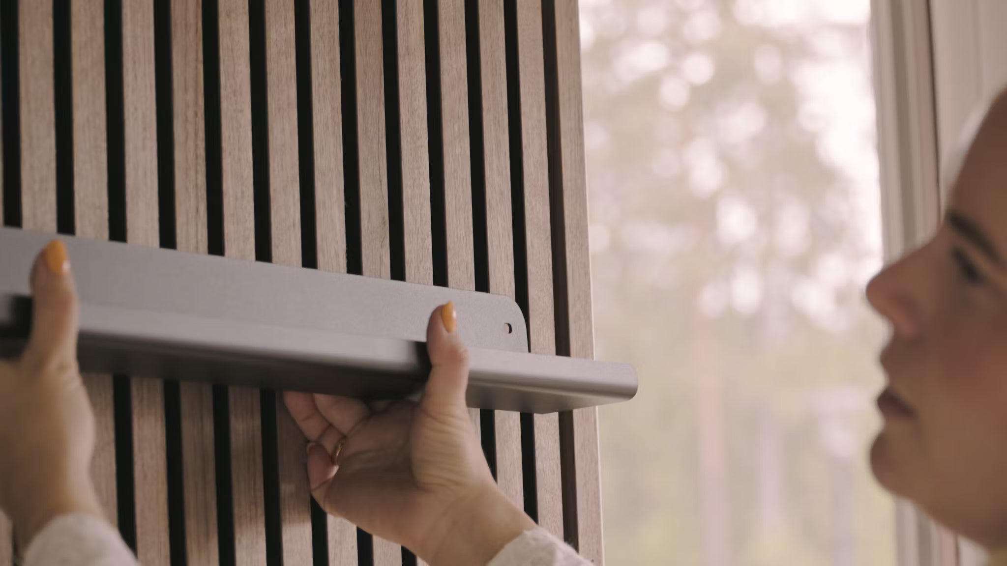 Una persona installa una mensola grigia su una parete di doghe acustiche Nordic Akupanel, accanto a una finestra con vista sugli alberi.