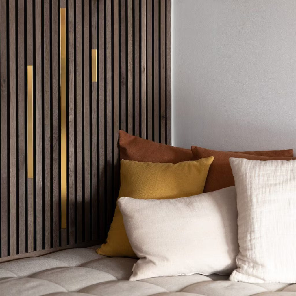Cuscini color crema, gialli e marroni su un divano beige con inserti, accanto a un moderno pannello acustico a doghe con accenti dorati, per un ambiente accogliente e contemporaneo.