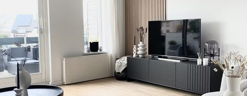 Soggiorno moderno con mobile TV nero, TV a schermo piatto, vasi decorativi e ampie finestre con luce naturale che si affacciano sul patio; Akupanel su una parete.