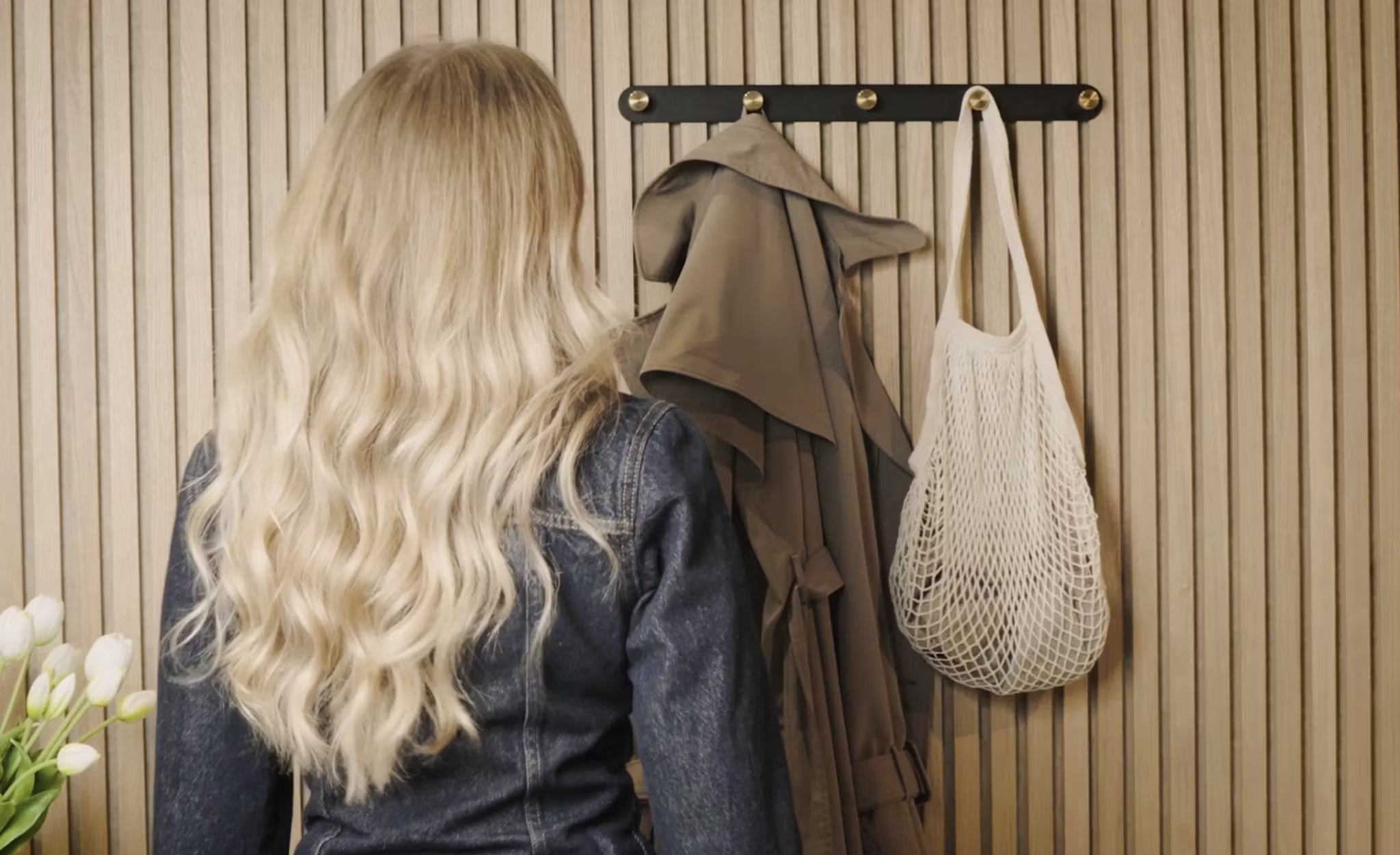 Una donna dai lunghi capelli biondi si trova di fronte a un appendiabiti con un trench, una borsa a rete bianca e tulipani bianchi freschi, accanto a un pannello di legno acustico Akupanel nellangolo.