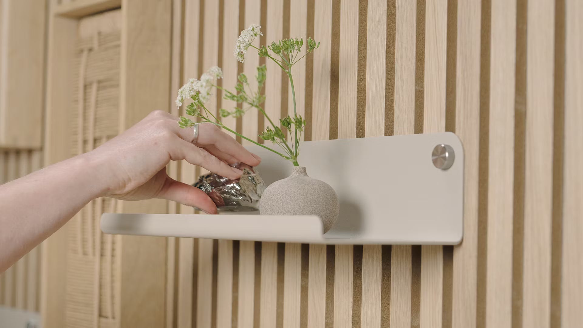 Una mano appoggia un piccolo oggetto decorativo accanto a un vaso di fiori bianchi su una mensola minimalista, appoggiata su un pannello a doghe in legno acustico nordico.