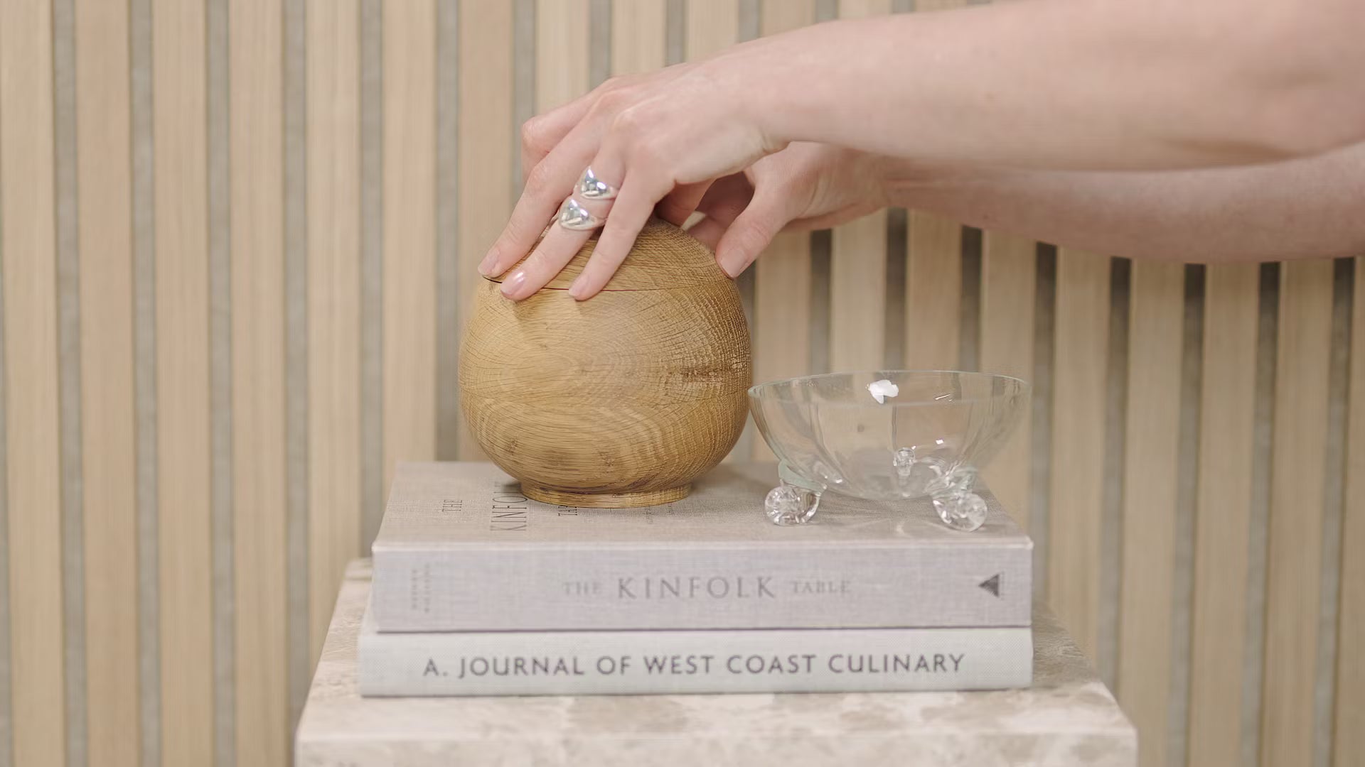 Una mano tocca una sfera di legno posta su libri impilati, tra cui Kinfolk Table e Journal of West Coast Culinary, accanto a una ciotola di vetro su un pannello acustico di doghe di legno.