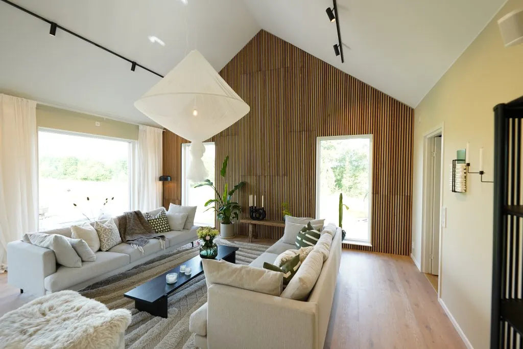 Soggiorno moderno con soffitto alto, ampie finestre, divani chiari, parete daccento in legno acustico Akupanel, piante da interno e abbondante luce naturale.
