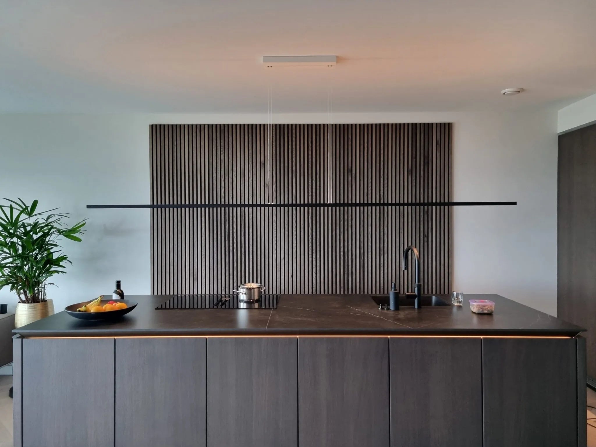 Cucina moderna con isola scura, lavello e fornelli a incasso, fruttiera e pianta in vaso. I pannelli verticali in legno acustico Akupanel accentuano la parete dietro lisola.