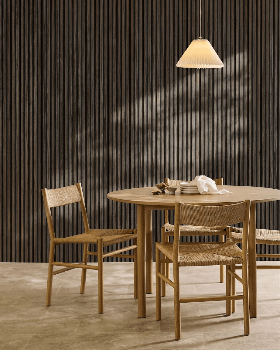 Un tavolo rotondo con quattro sedie intrecciate è disposto accanto a un pannello WoodUpp Akupanel Black Oak Pannello acustico, sotto una lampada a sospensione plissettata.