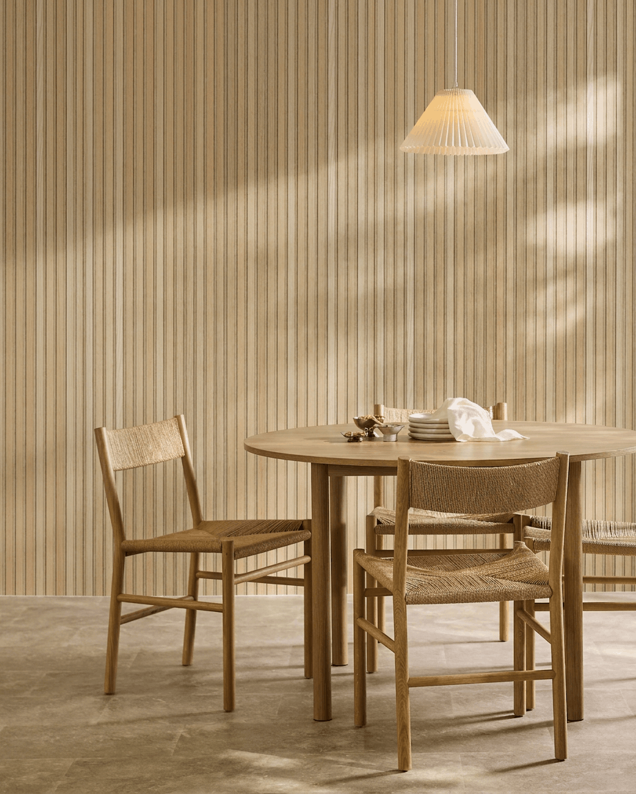 Zona pranzo minimalista con tavolo rotondo, sedie intrecciate e lampada a sospensione accanto a un pannello acustico in legno WoodUpp Akupanel in Classic Rovere con feltro beige.