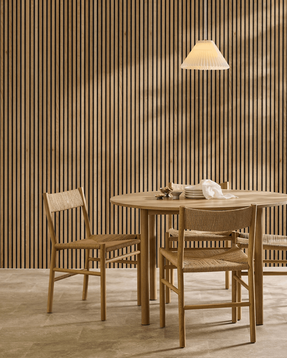 Zona pranzo minimalista con un tavolo in legno, due sedie e un pannello acustico in legno WoodUpp Akupanel in Rustic Brown Rovere (300x60 cm), illuminata da una lampada a sospensione.