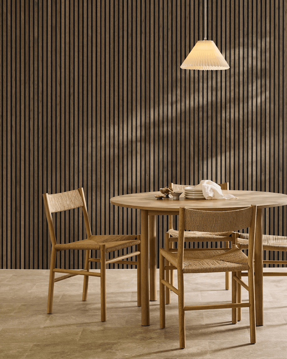 Zona pranzo minimalista con tavolo rotondo, sedie intrecciate e lampada a sospensione, con un pannello fonoassorbente a doghe WoodUpp Akupanel Rustic Grey Oak.