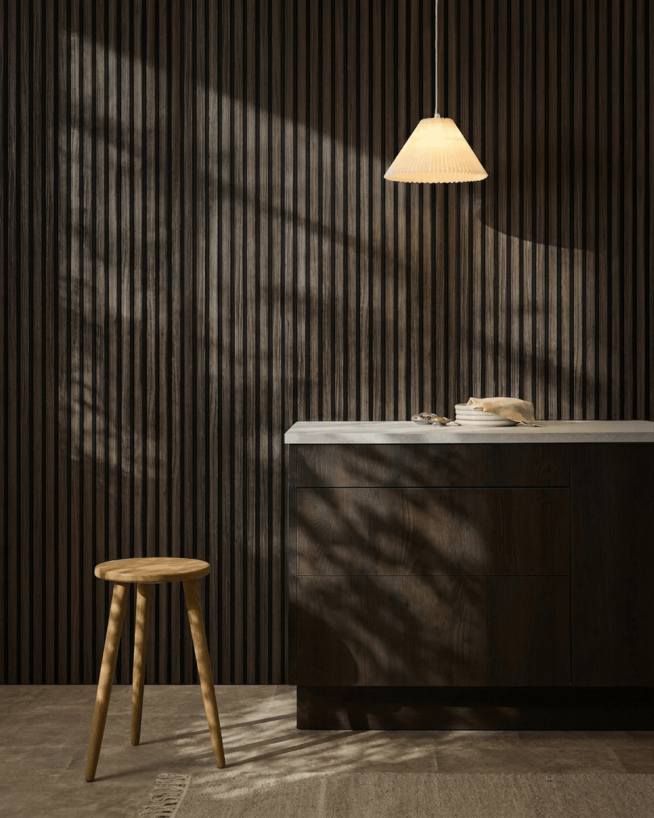 Cucina minimalista con pannelli acustici WoodUpp Akupanel Black Oak sulla parete, sgabello in legno, piano di lavoro bianco, lampada a sospensione beige e ombre illuminate dal sole.