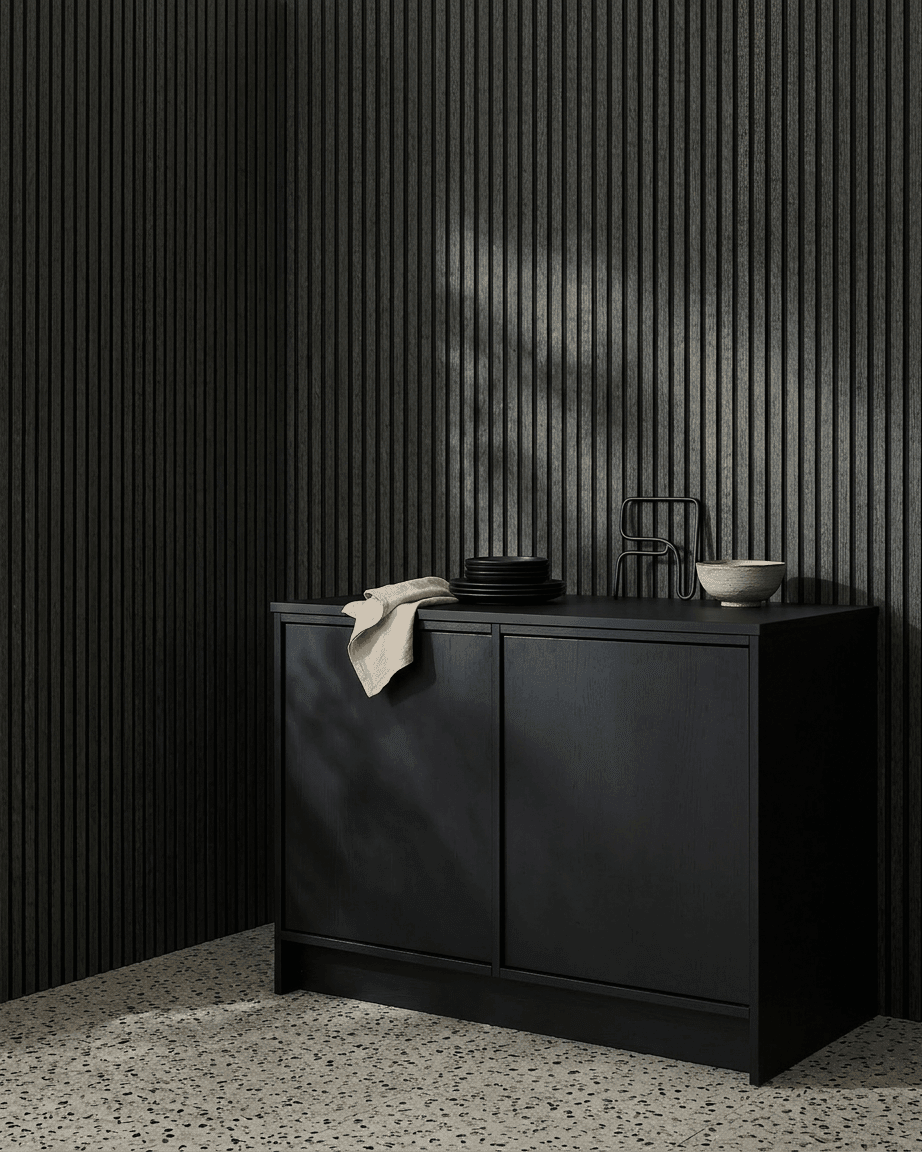 Un mobile black accanto a un pannello acustico in legno WoodUpp Akupanel in Charcoal/Black Felt, con sopra ciotole e piatti, su un pavimento chiaro in terrazzo.
