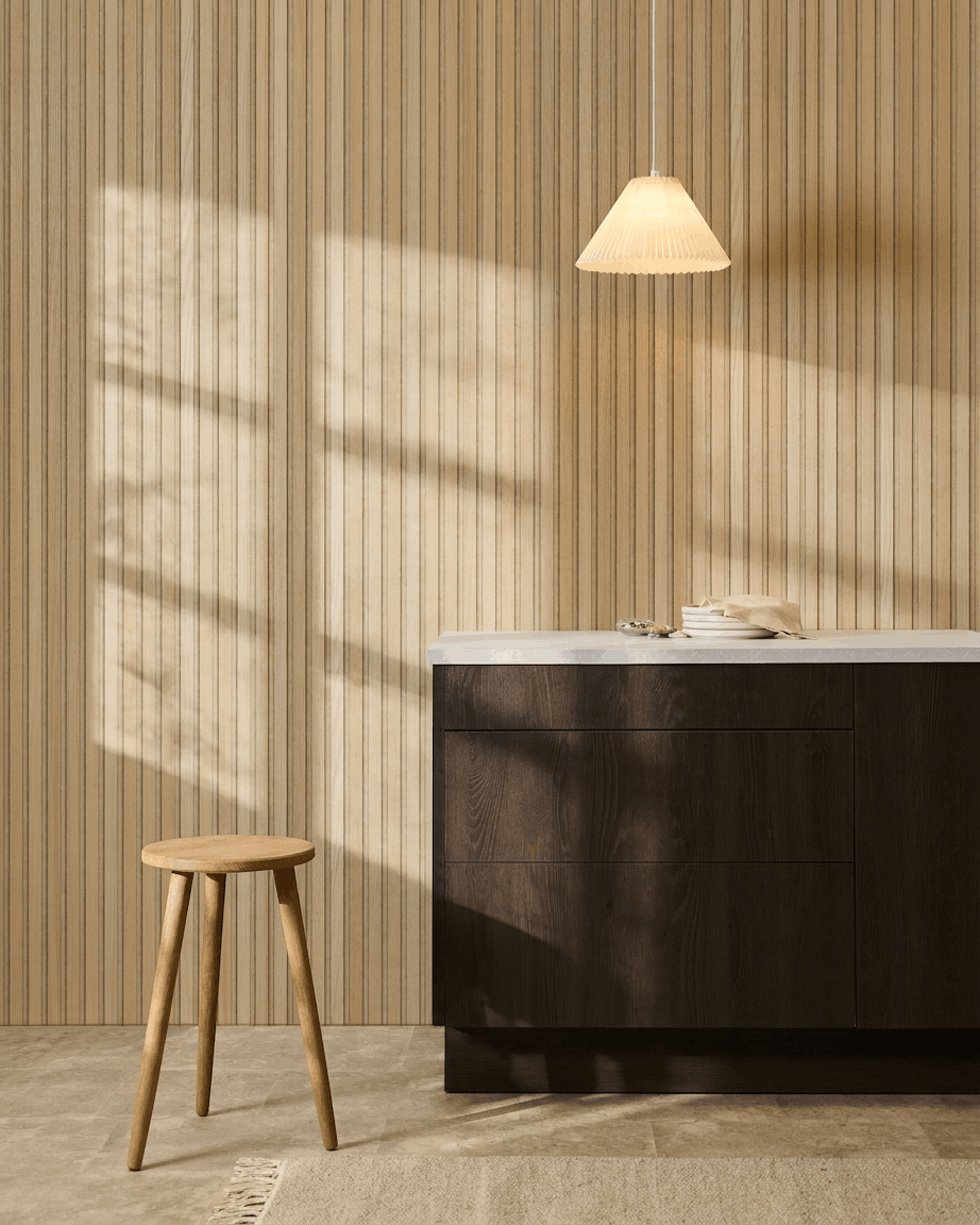 Cucina scandinava con pannello acustico a doghe WoodUpp Akupanel Classic Rovere, mobili in legno scuro, sgabello in legno, lampada a sospensione e luce del sole che proietta ombre.
