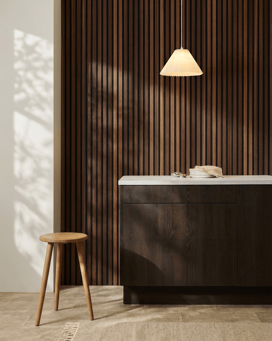 Interno minimalista con uno sgabello in legno, un mobile in legno scuro, una lampada a sospensione e un pannello acustico in legno a doghe WoodUpp Akupanel in Natural Wood/termotrattato Larch.