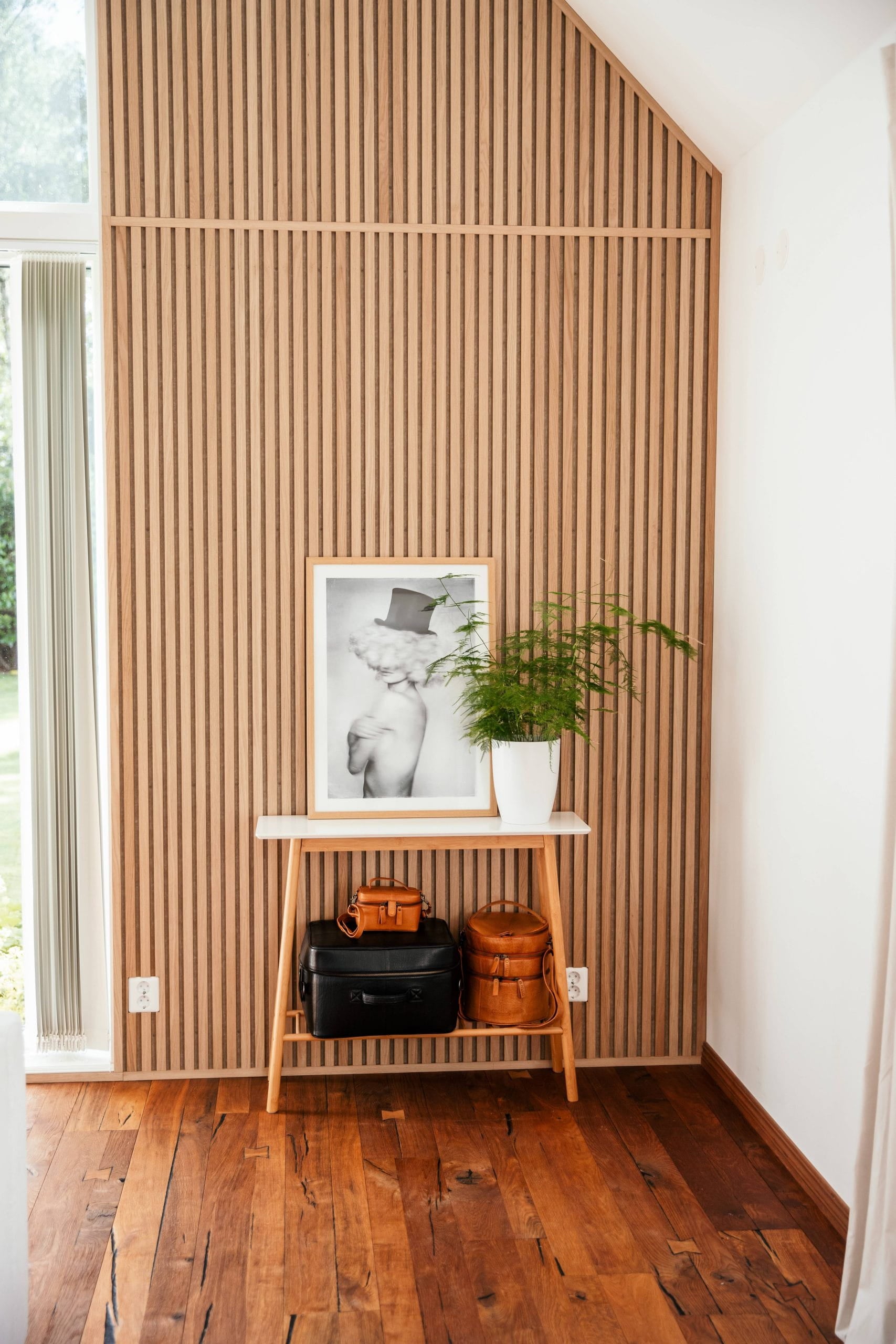 Un tavolino di legno con borse, una foto incorniciata e una pianta in vaso è appoggiato a un pannello acustico nordico in una stanza luminosa con pavimenti in legno.