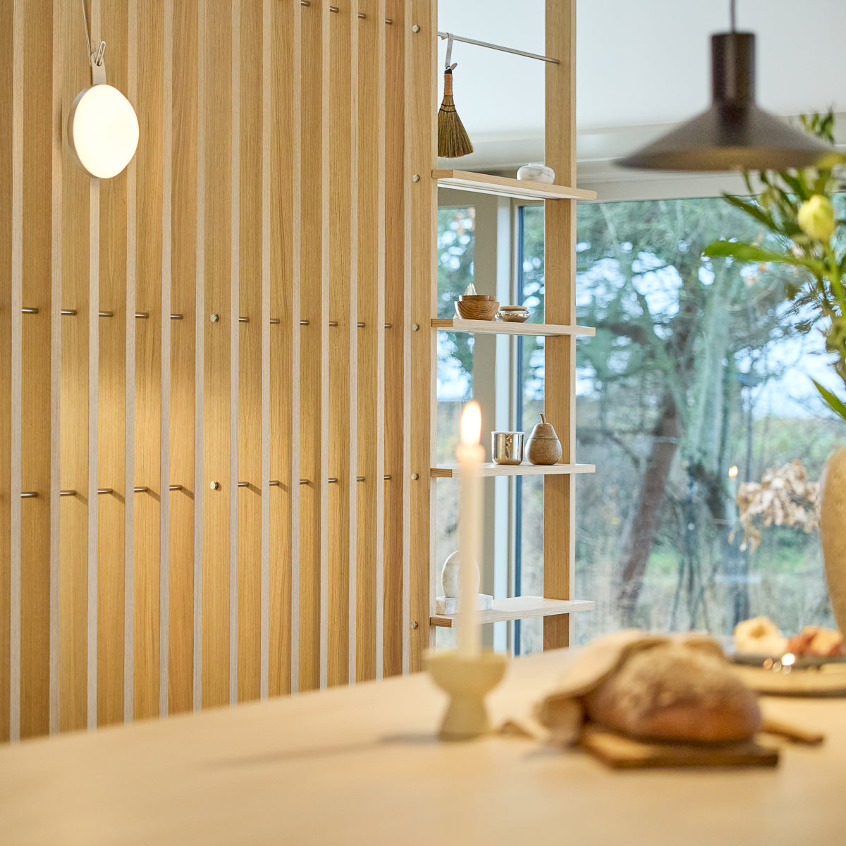 Interno minimalista con un divisorio per ambienti in legno Nordic EMBRACE, candela accesa, pane fresco sul tavolo, decorazioni curate sugli scaffali e finestra che incornicia alberi all'esterno.