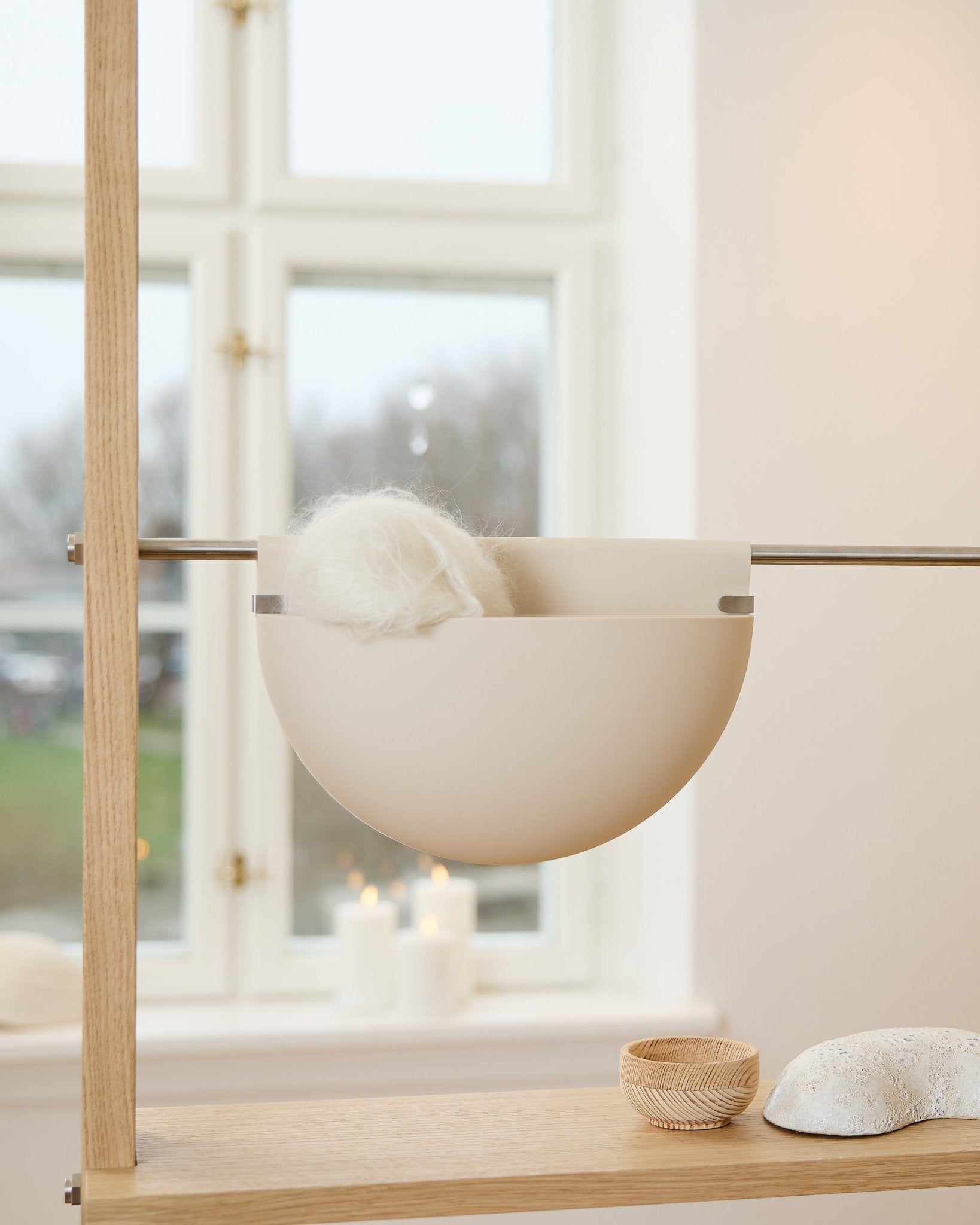 Una ciotola bianca con un oggetto soffice poggia su una mensola in legno vicino a candele, decorazioni e a un accessorio acustico WoodUpp Gro Soft Pocket BM2 in beige chiaro accanto alla finestra.