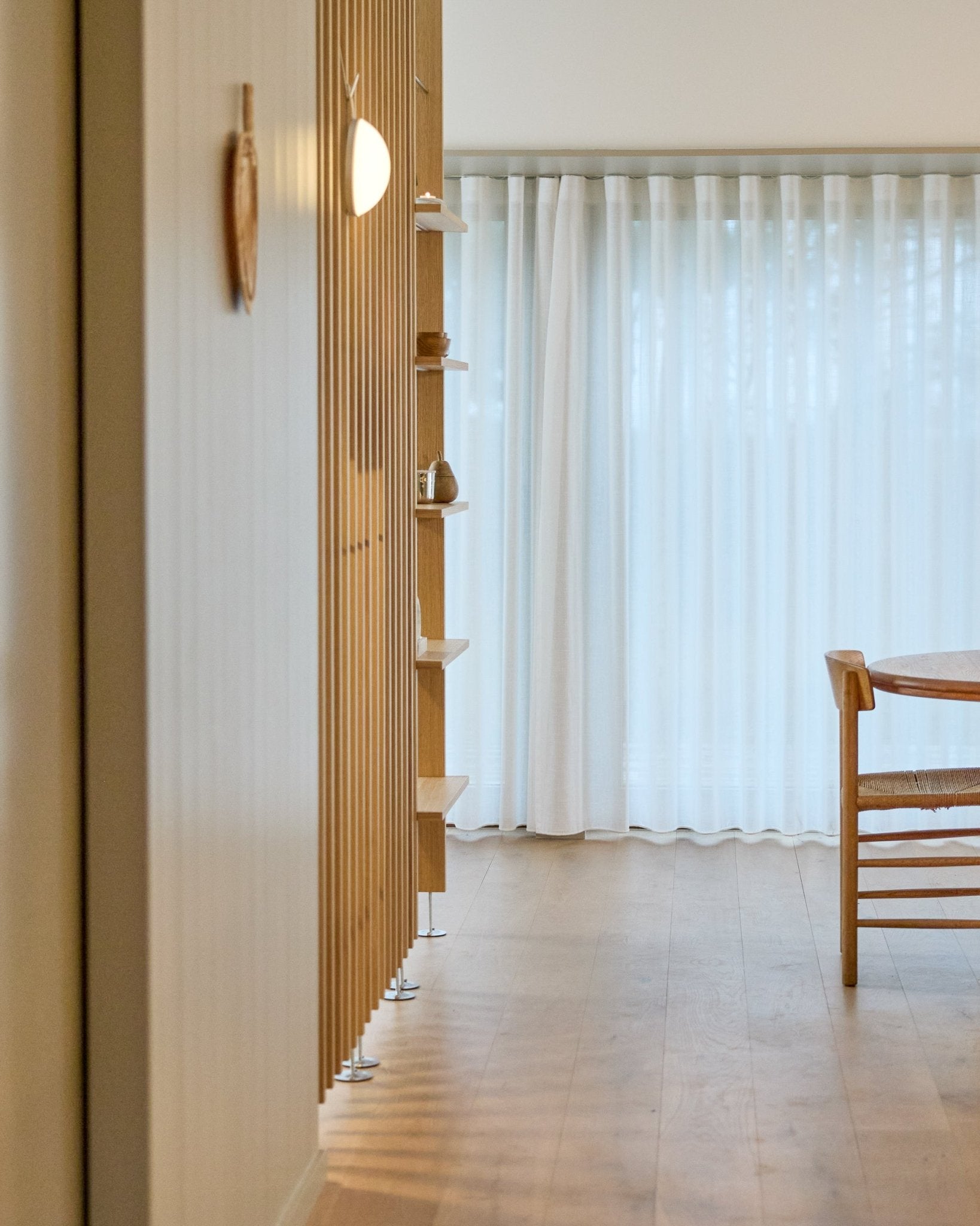 Ambiente minimalista con un divisorio per ambienti WoodUpp Embrace da pavimento a soffitto, pavimento in legno, mensole, sedia e tende velate davanti a finestre large.
