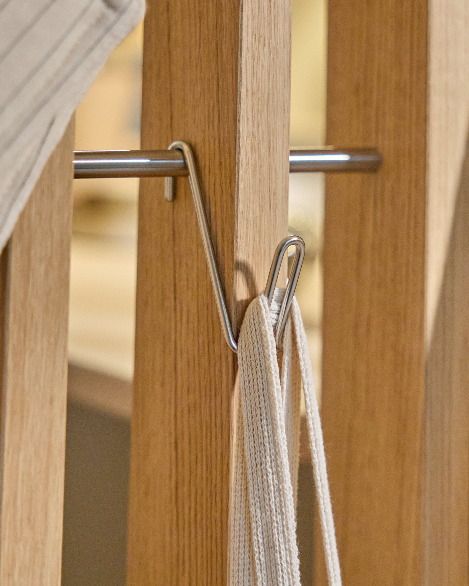 L’Alva Wire Hook / BM1 (acciaio inox Steel, Short) di WoodUpp è montato su una guida in legno Pannello acustico e sostiene una cinghia bianca con asta in metallo; sfondo beige.