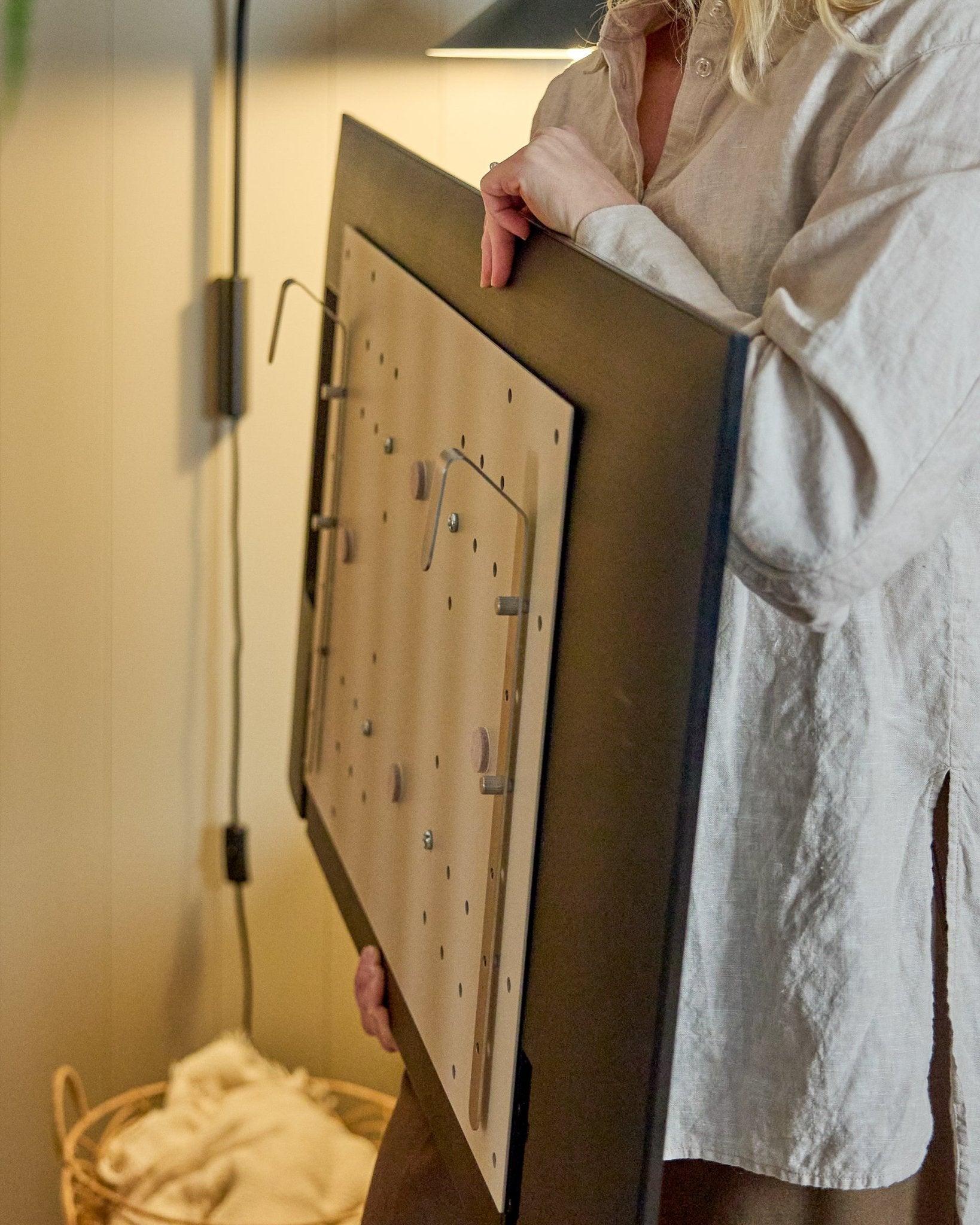 Una persona con una camicia chiara tiene in mano il WoodUpp Nell BM1 TV-Mount in beige, 64x44 cm, compatibile con i pannelli acustici in legno Nordic, per installazioni senza parete.
