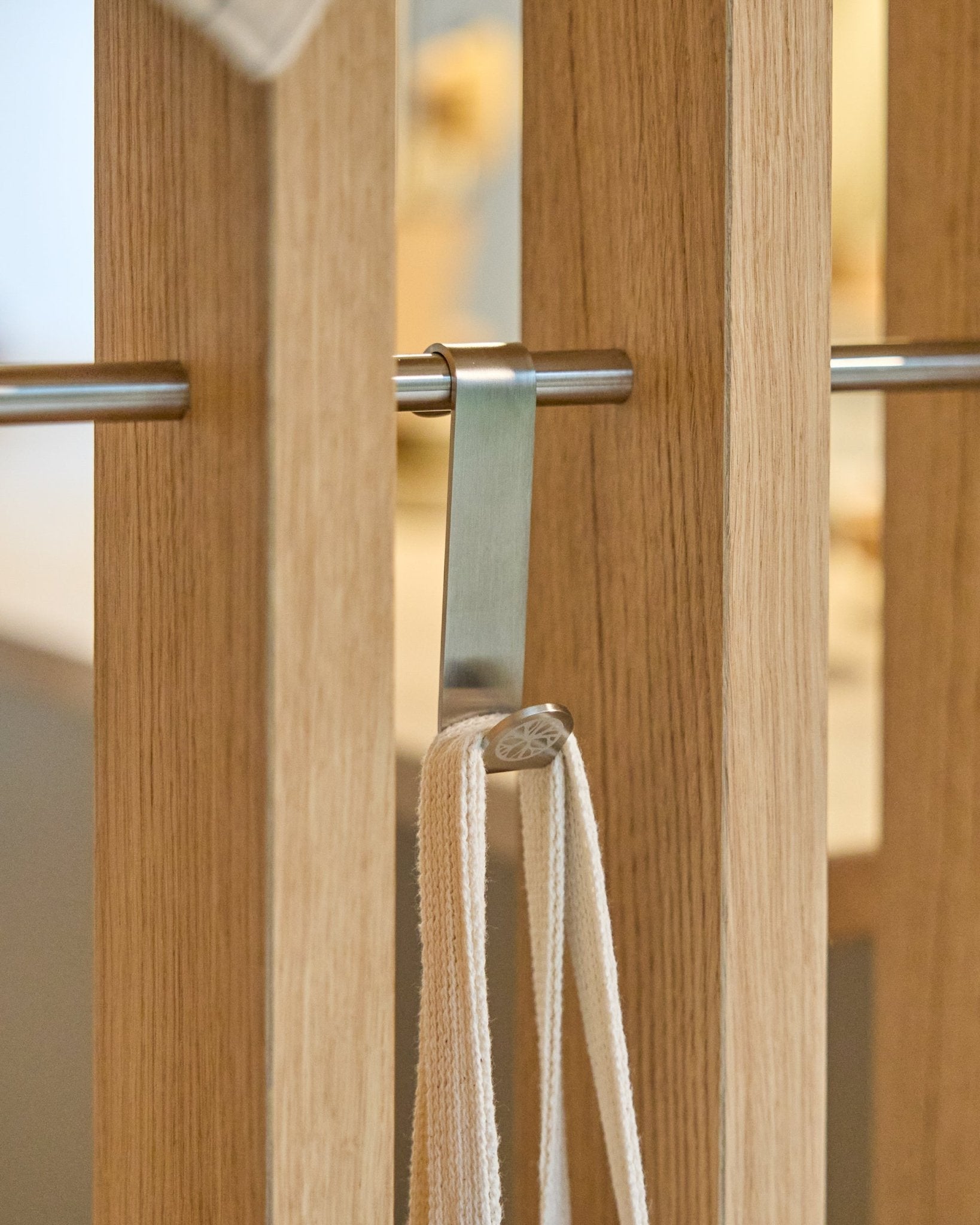 Un gancio Alva S-Hook WoodUpp in steel inox fissa una cinghia beige a una barra montata tra pannelli acustici in legno a doghe vertical. Il set include 2 ganci.