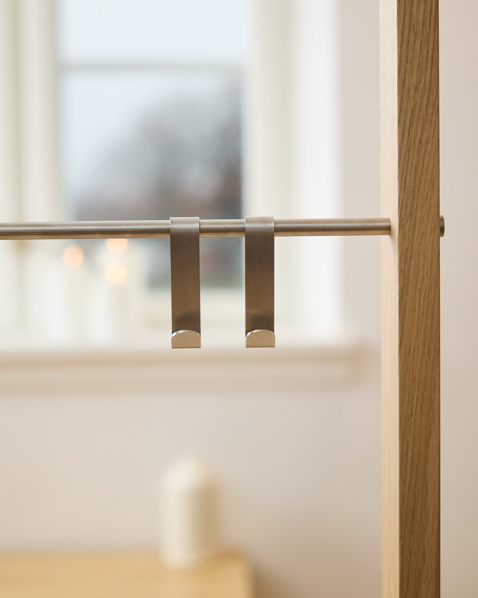 WoodUpp Alva S-Hook BM1  BM2 (2 pz) in steel inox, appesi a una barra per pannello acustico in legno; finestra sfocata e candele accese sullo sfondo.
