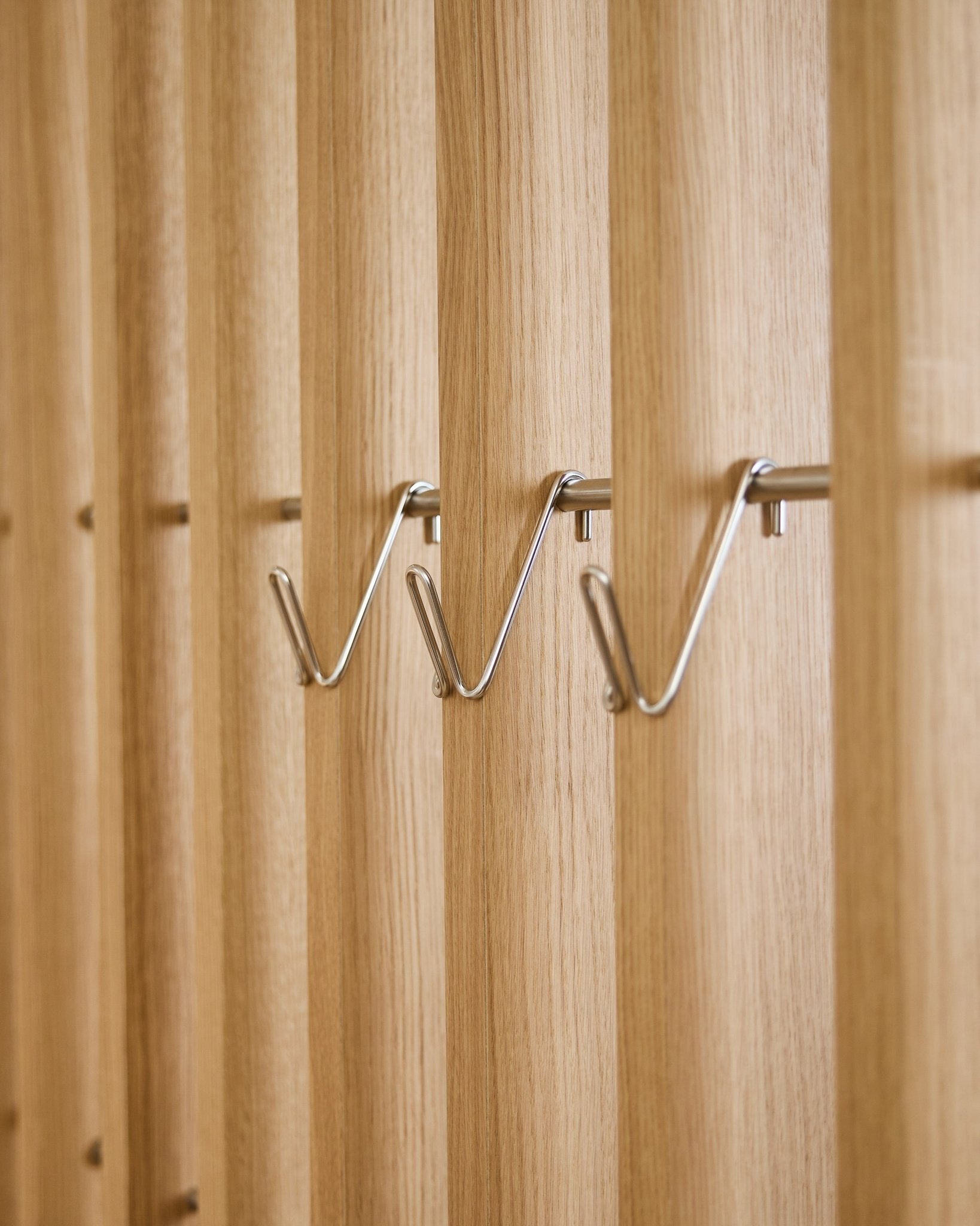 Tre accessori WoodUpp Alva Wire Hook / BM1 corti in acciaio inox steel sono montati in modo uniforme in fila su un pannello acustico in legno a doghe vertical.