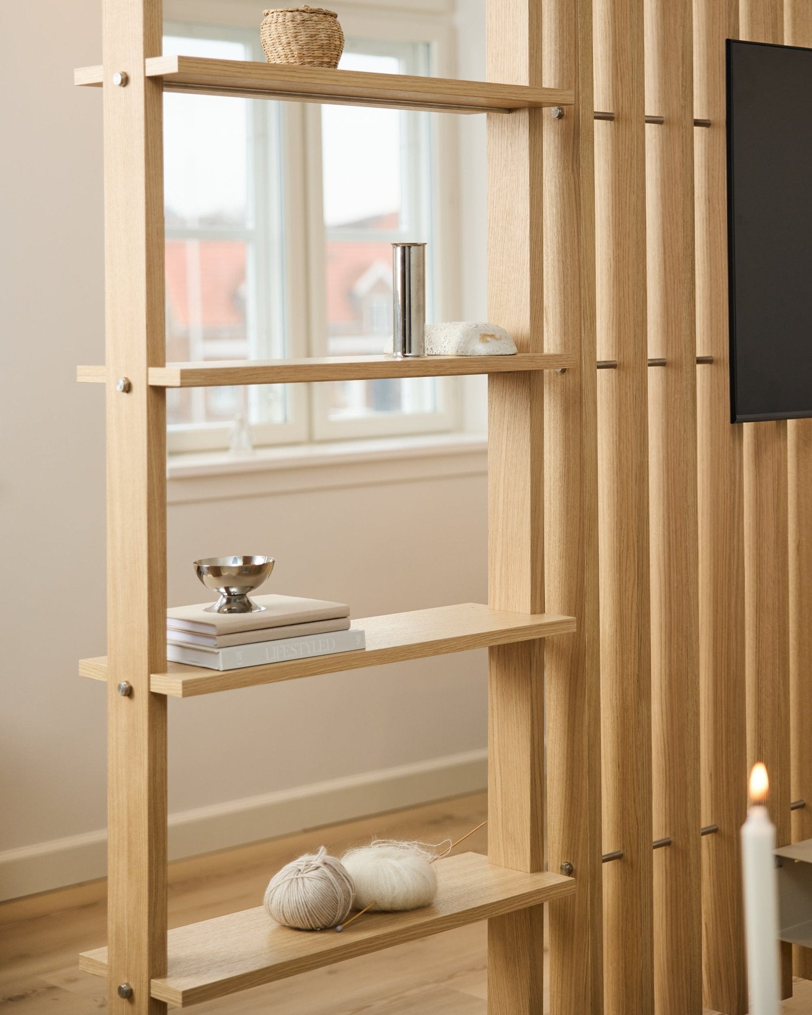 Separé in legno chiaro Rovere di WoodUpp, con doghe vertical e mensole che ospitano libri e gomitoli di lana.