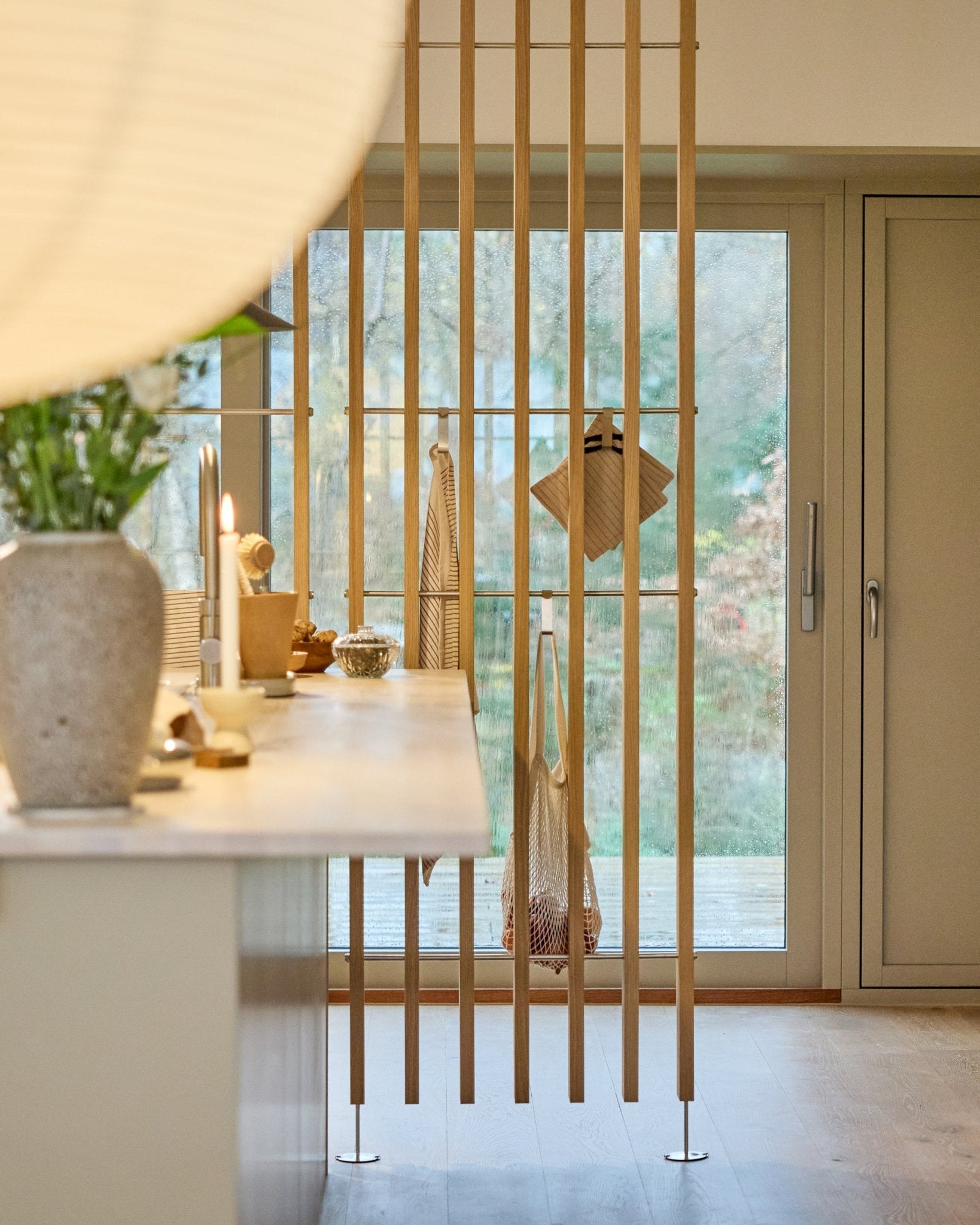 Cucina minimalista con un divisorio WoodUpp Embrace (BM1, 286-302 cm) in impiallacciatura Rovere, con piani di lavoro chiari, un vaso con fiori e vista sul giardino.
