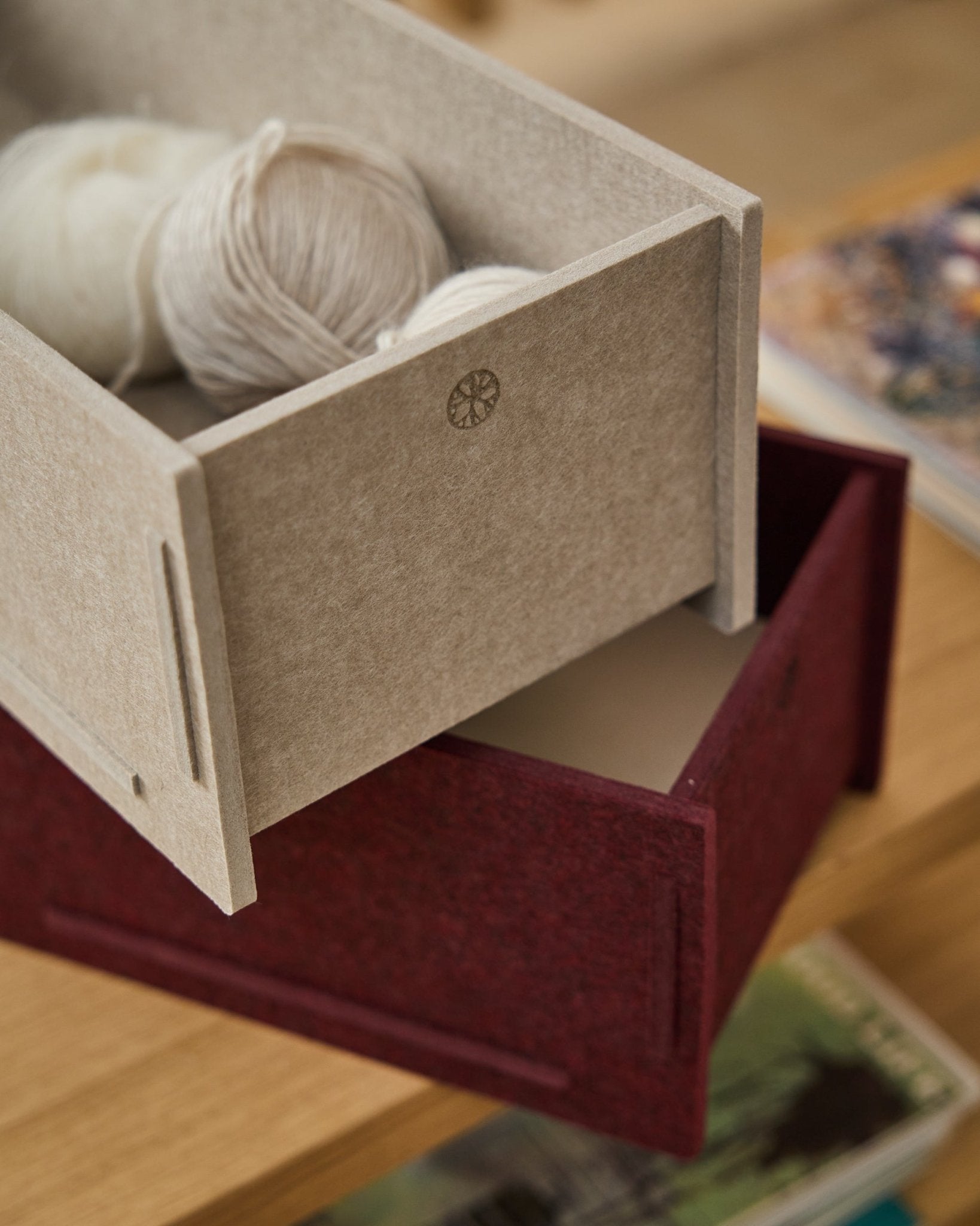 Un contenitore in feltro PET beige pieno di filati è appoggiato sopra una WoodUpp Vigg Felt Box/BM2 chiusa in ox red, su un pavimento in legno con libri sullo sfondo.