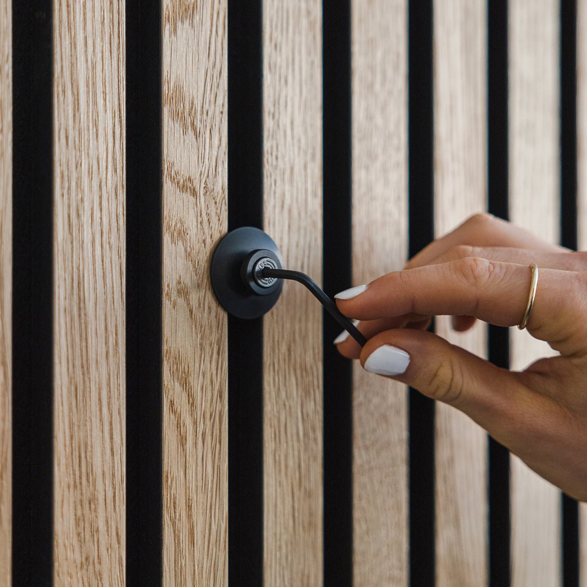 Una mano con smalto bianco ruota un gancio in alluminio black | Heavy Dupps di WoodUpp su un pannello acustico in legno vertical Akupanel.