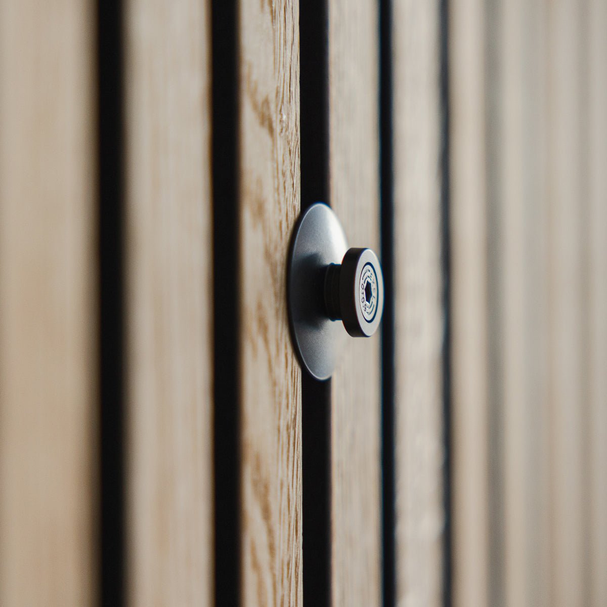 Primo piano dello Dupps | Spioncino Heavy di WoodUpp in alluminio black, installato su una porta in legno scanalata con doghe acustiche Akupanel, con la lente a fuoco.
