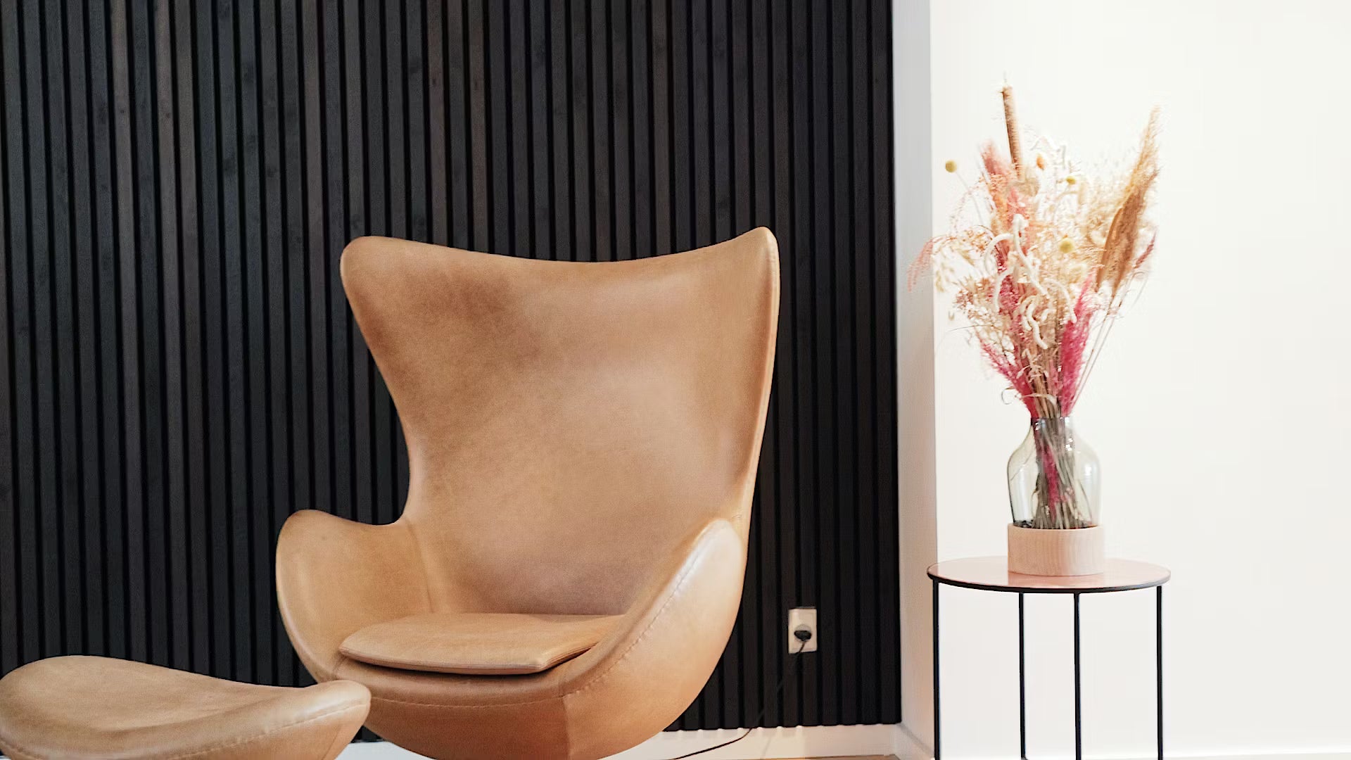 Sedia e pouf in pelle abbronzata accanto a un tavolino con fiori secchi, su un pannello acustico nero a doghe di legno e uno sfondo bianco.