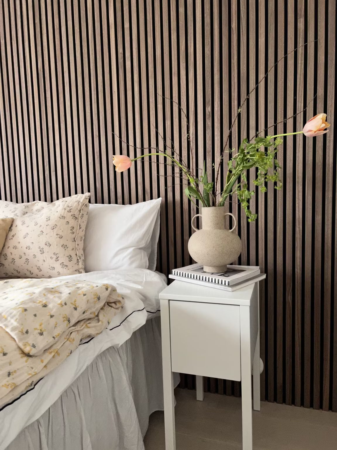 Una camera da letto con un letto bianco con biancheria floreale e un vaso di fiori, con una parete daccento in legno nordico a doghe acustiche Akupanel dietro il letto.