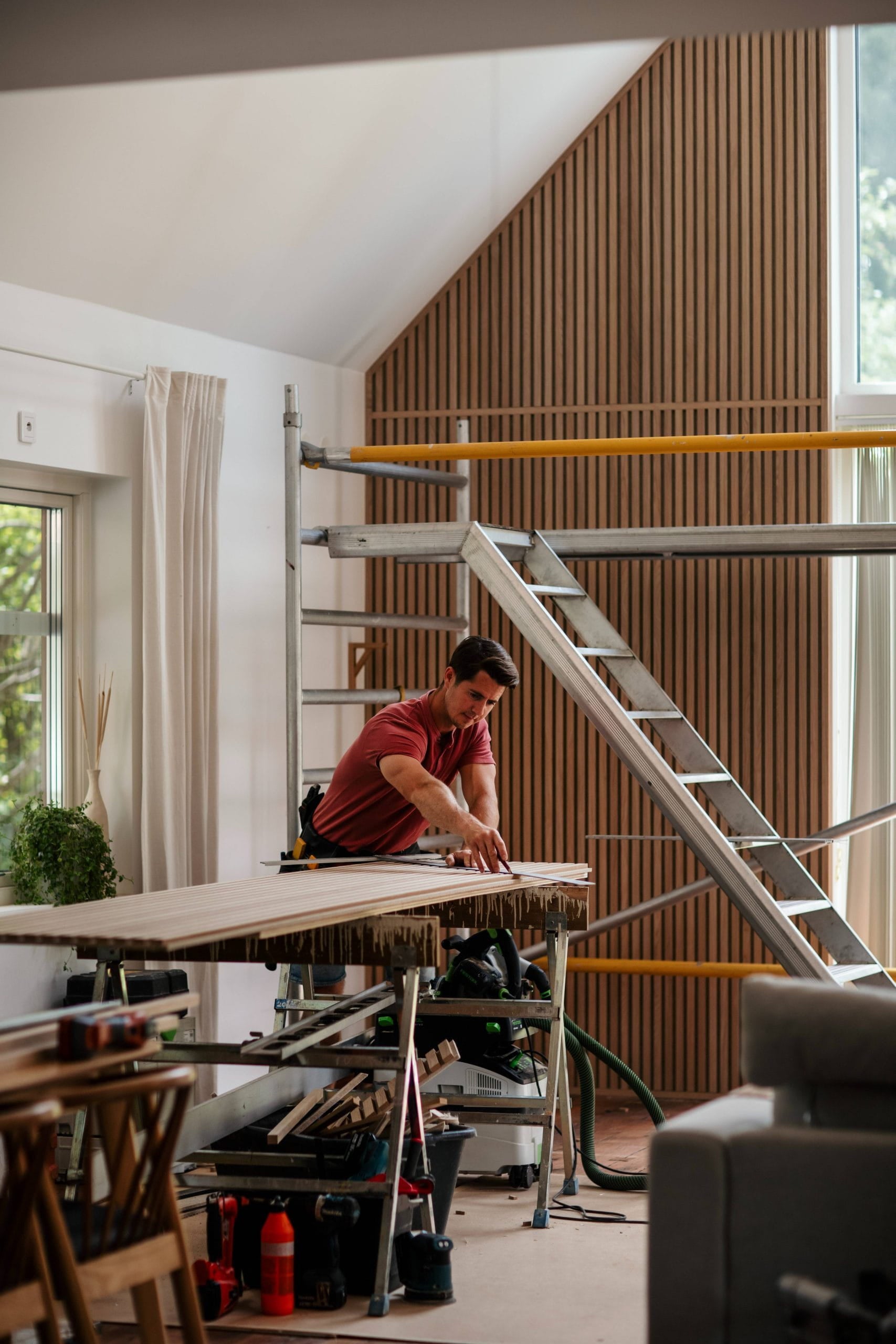 Un uomo misura una tavola di legno su un banco di lavoro in una stanza moderna, parzialmente ristrutturata, con impalcature e grandi finestre, che suggeriscono linstallazione di pannelli acustici.