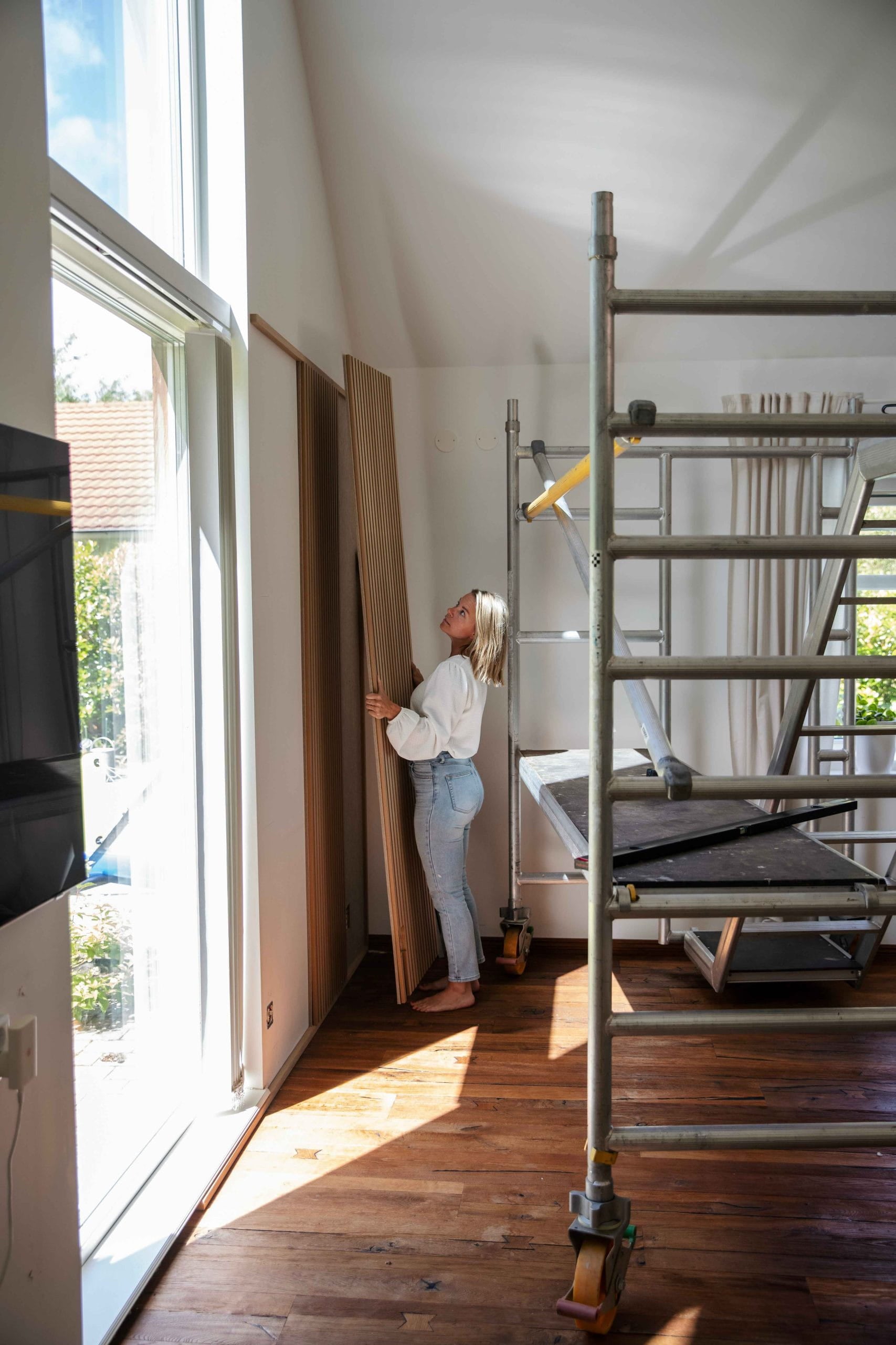 Una donna si trova in una stanza illuminata dal sole con grandi finestre e impalcature, tenendo in mano un pannello di legno acustico Akupanel su un pavimento di legno lucido.