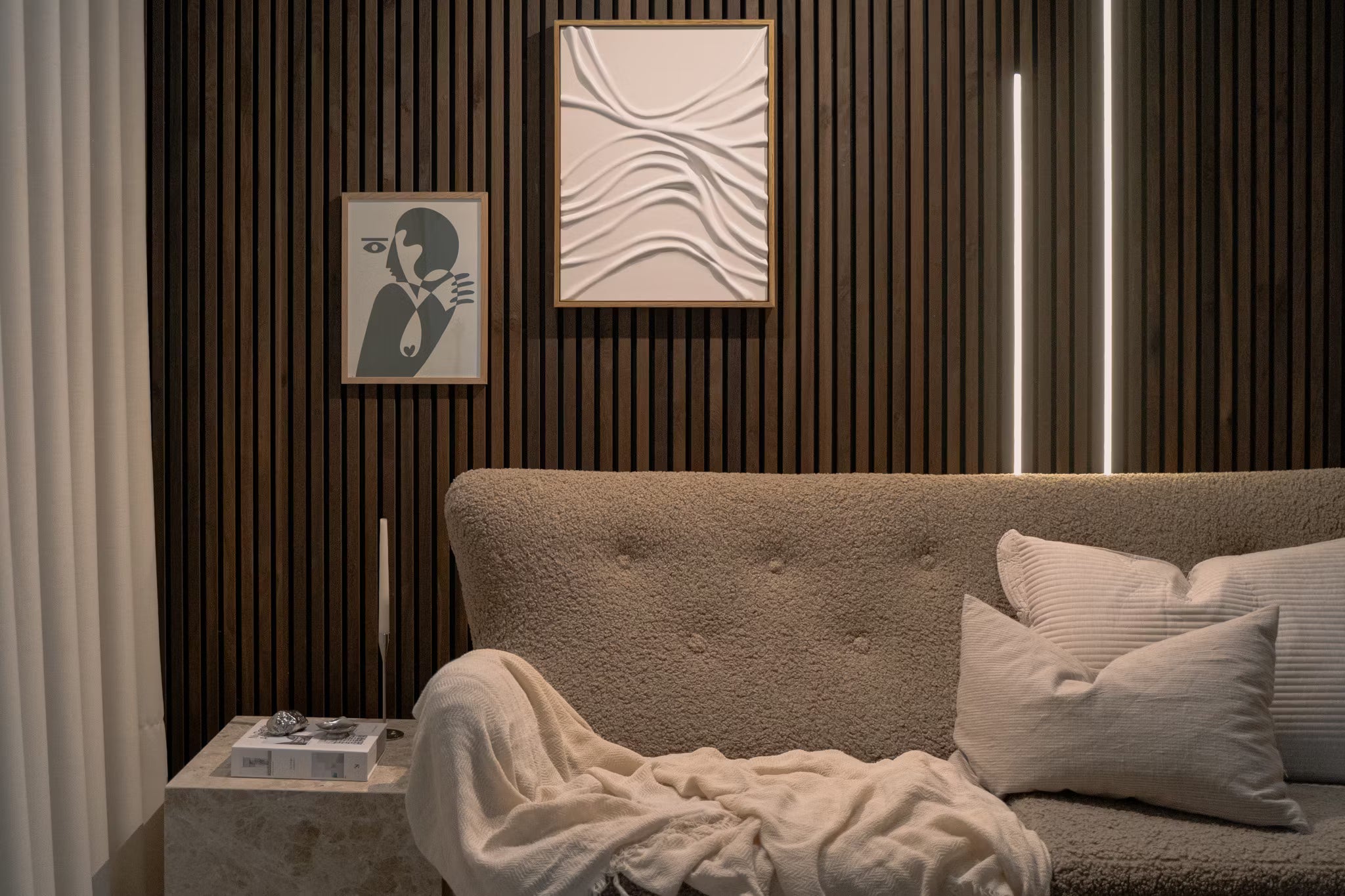Soggiorno moderno con divano beige testurizzato, cuscini bianchi, pannelli acustici astratti e parete verticale in doghe di legno Akupanel con illuminazione dambiente soffusa.