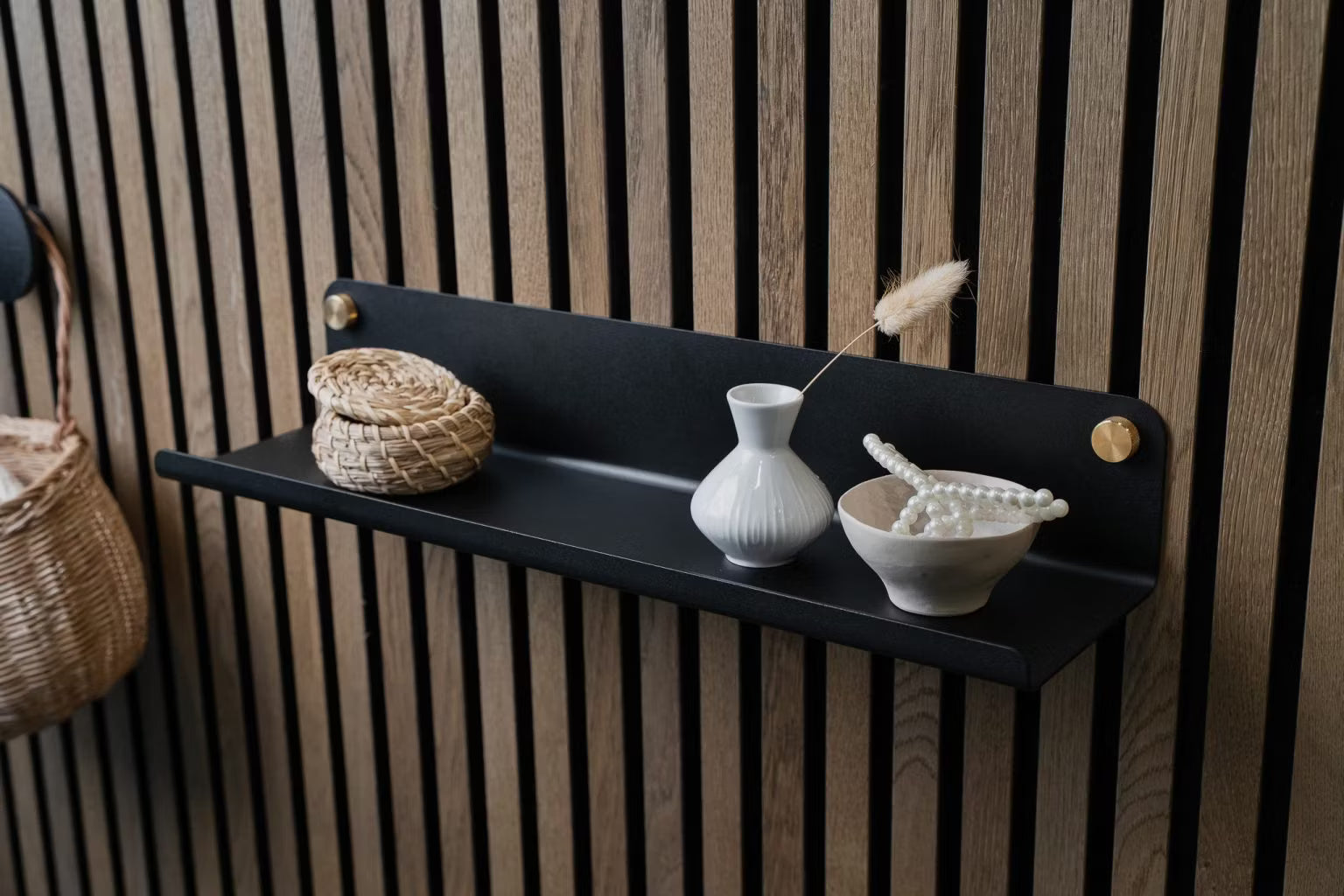Una mensola a muro nera con un piccolo cesto di vimini, un vaso bianco e una ciotola di perle in ceramica montati su un pannello acustico a doghe in legno nordico.