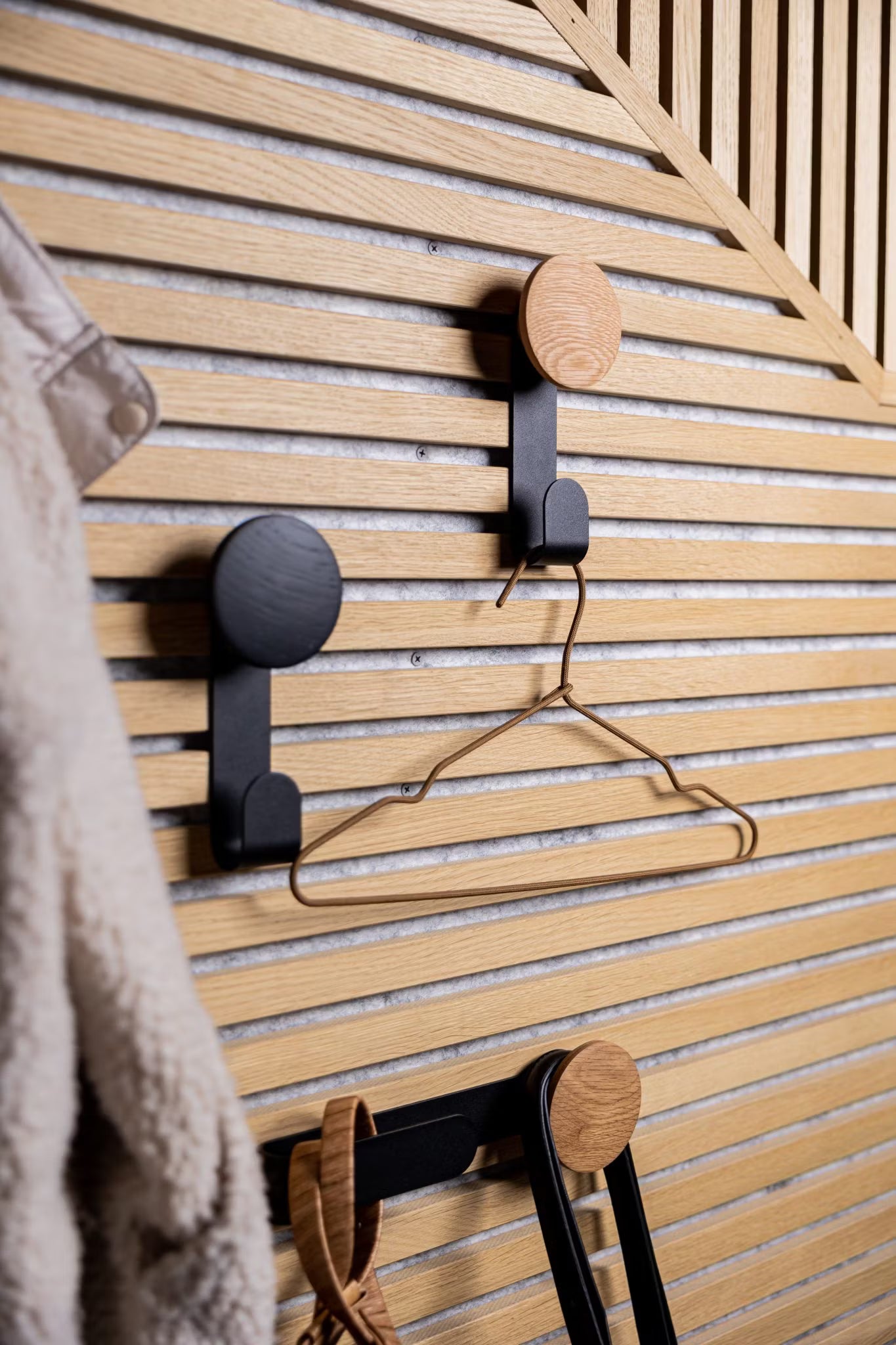 Tre ganci moderni montati su una parete a doghe in legno acustico Akupanel in stile nordico ospitano una gruccia di metallo e accessori, con un cappotto sfocato parzialmente visibile sulla sinistra.