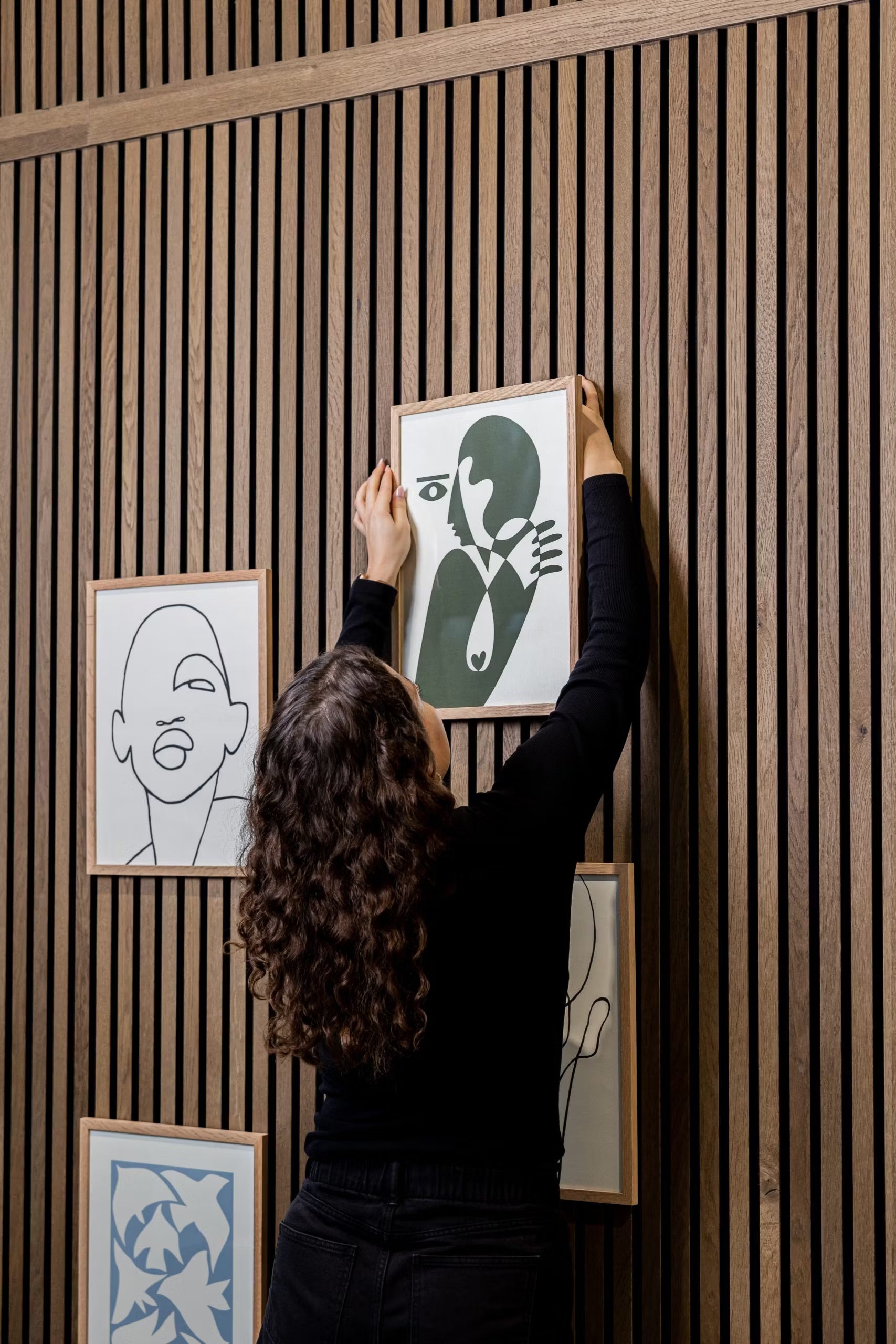 Una persona con i capelli ricci appende un accessorio WoodUpp® CREATE DUPPs a un pannello acustico a doghe in legno, circondata da arte minimalista e arredi moderni.